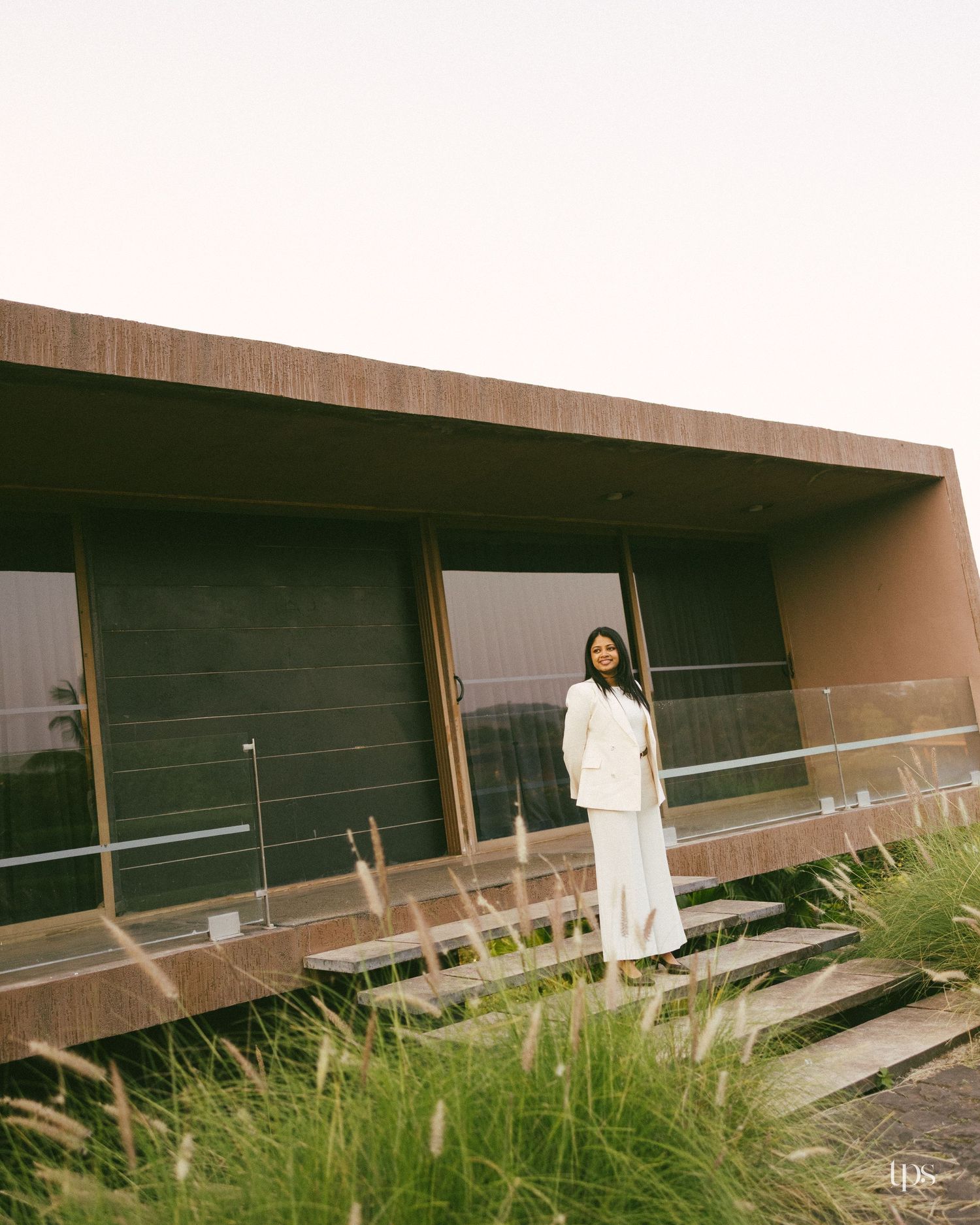 Woman in a white blazer and long skirt stands on wooden steps beside a modern brown building with large windows, surrounded by tall grasses.