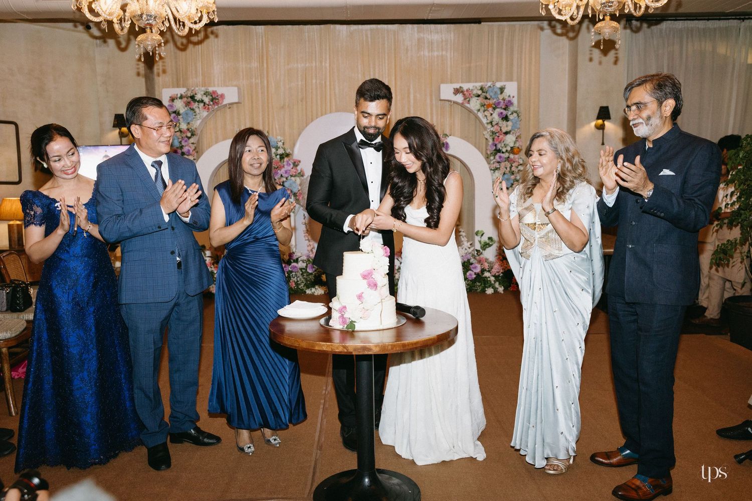 Bride and groom cut their wedding cake together as smiling guests in formal attire applaud in an elegant reception hall with floral decor.