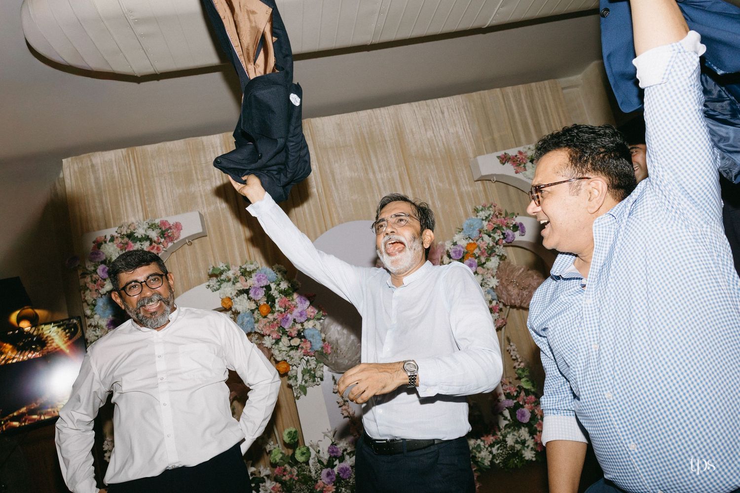Three men in white and light blue shirts laugh and celebrate at an indoor event, one tossing a jacket in the air beside floral decorations.