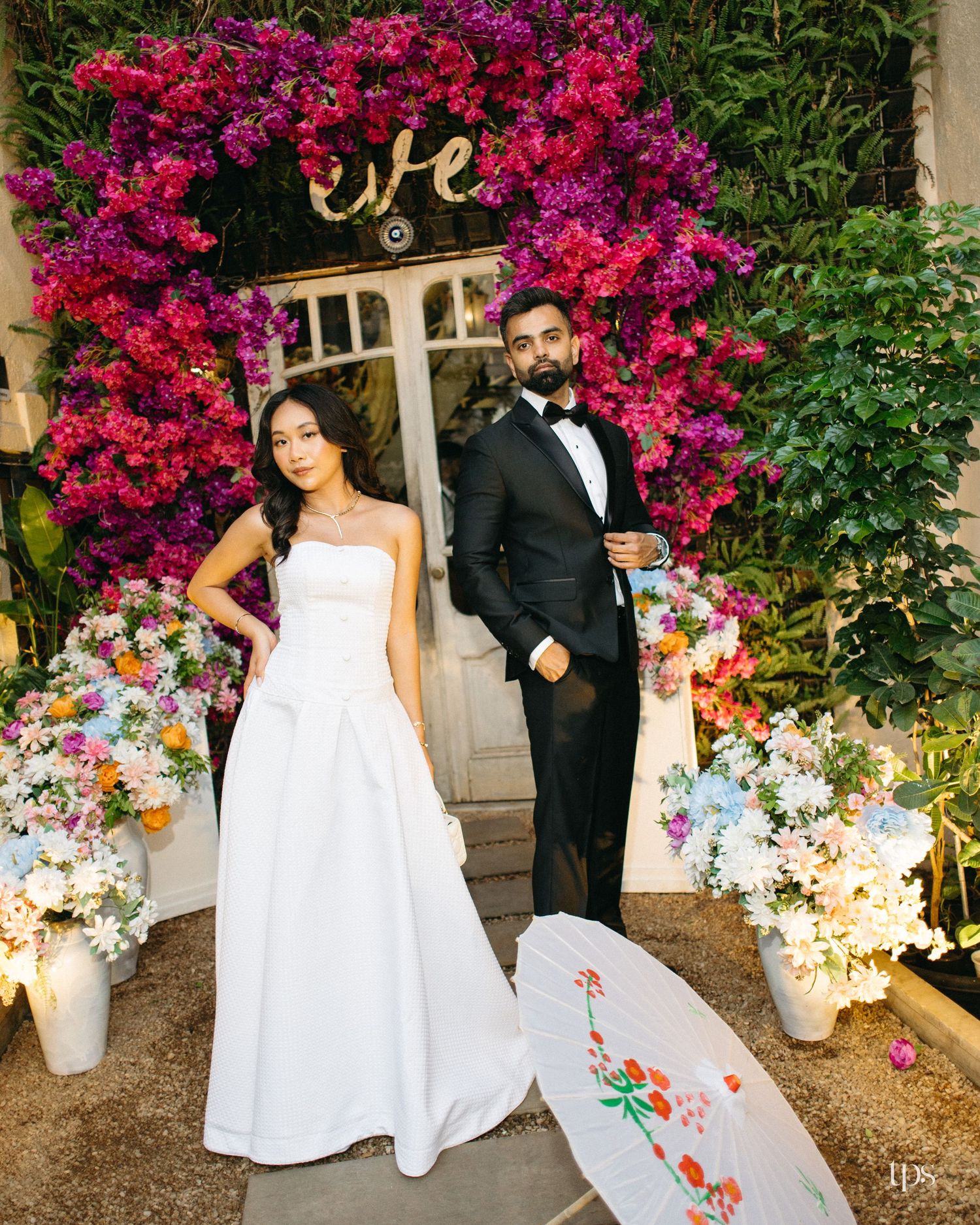 Bride in a white strapless gown and groom in a black tuxedo pose before a doorway framed by pink and purple bougainvillea at a wedding.