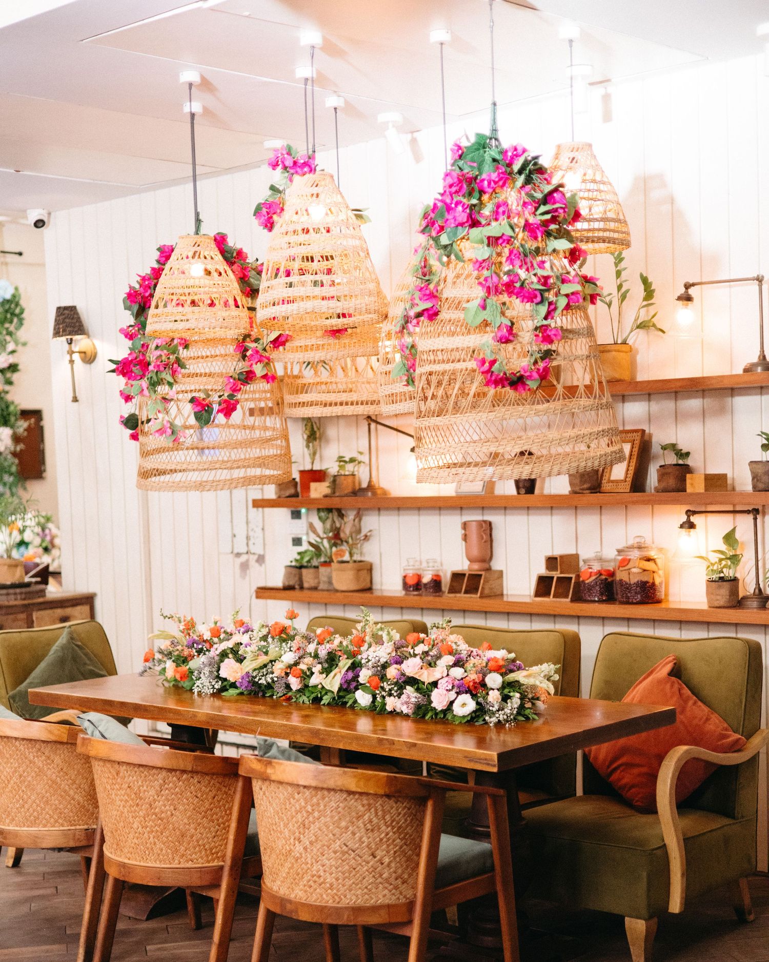 Cozy restaurant interior with a wooden table, pink floral centerpiece, rattan chairs, woven pendant lamps and green armchairs.