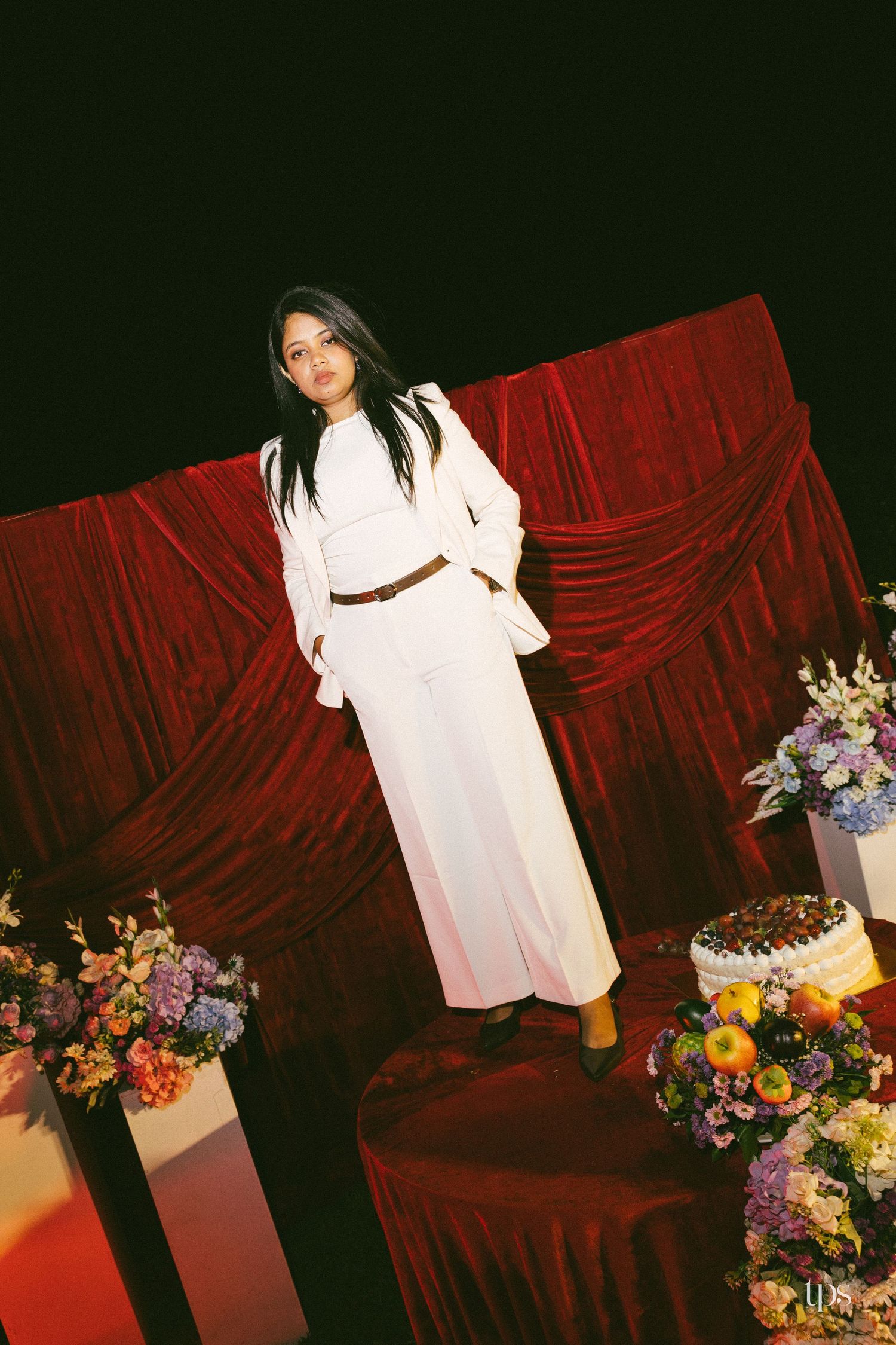 Woman in a white blazer, wide-leg pants and brown belt stands on a red velvet stage with floral arrangements and a decorated cake.