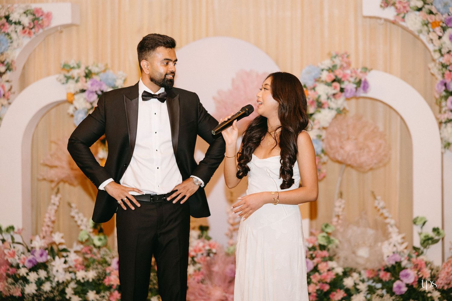 Groom in black tuxedo and bow tie smiles as a woman in a white dress speaks into a microphone on a stage decorated with pastel flowers.