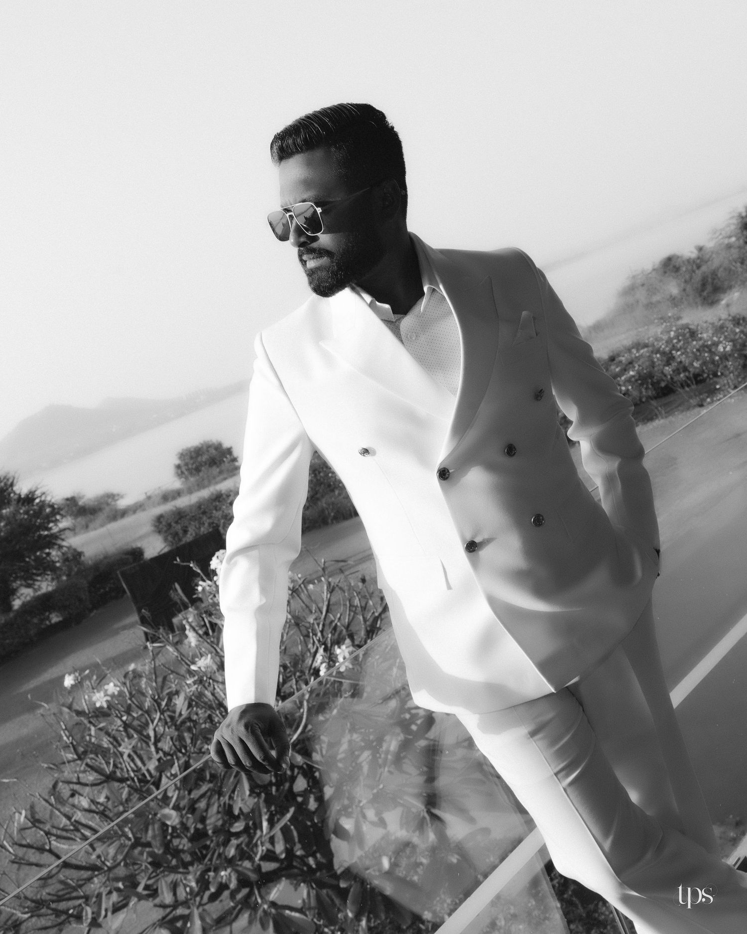Black and white photo of a bearded man in sunglasses and a double-breasted white suit leaning on a railing beside plants with a lake behind.