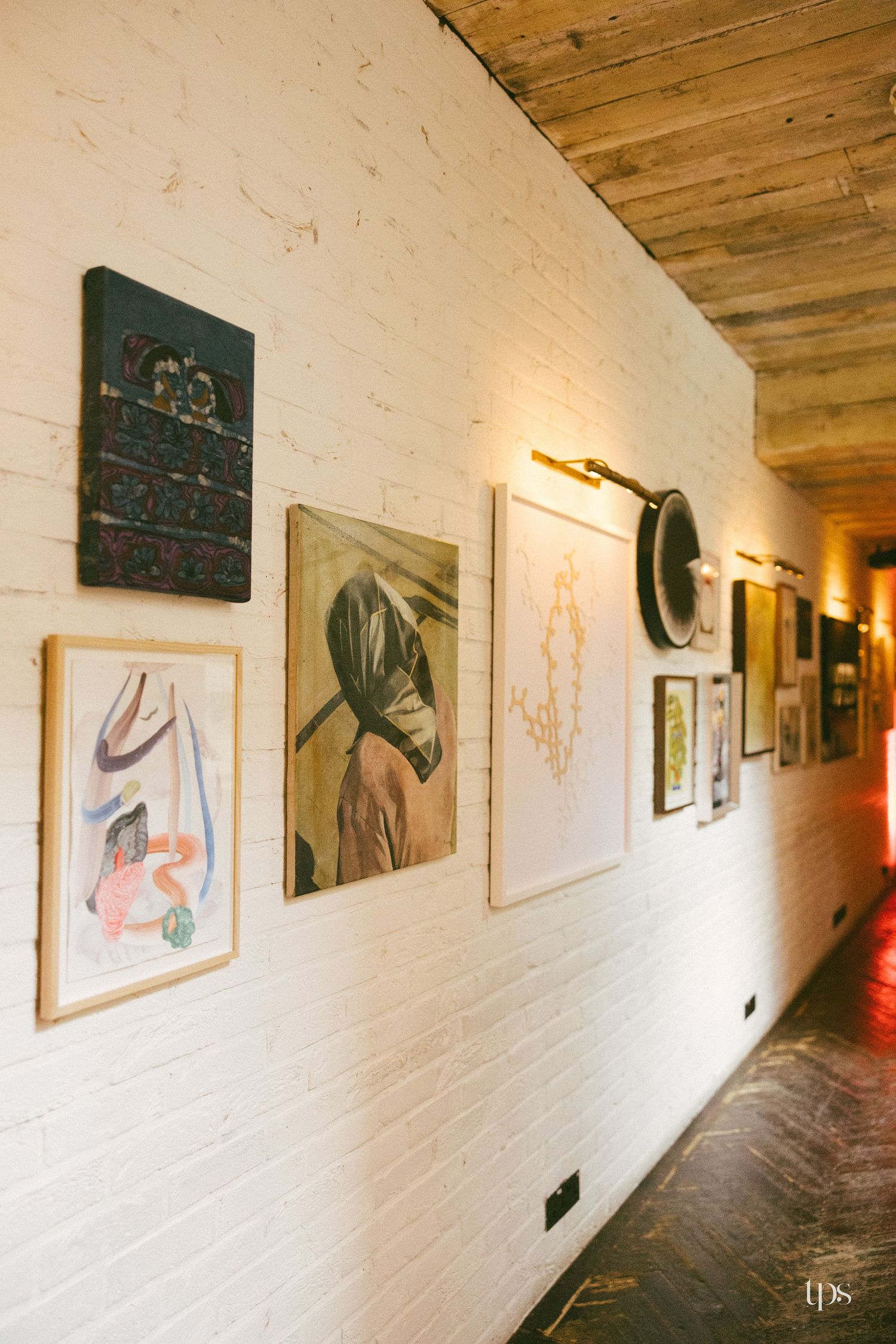A white brick hallway gallery wall displaying eclectic framed artwork under warm lighting with a wooden ceiling.
