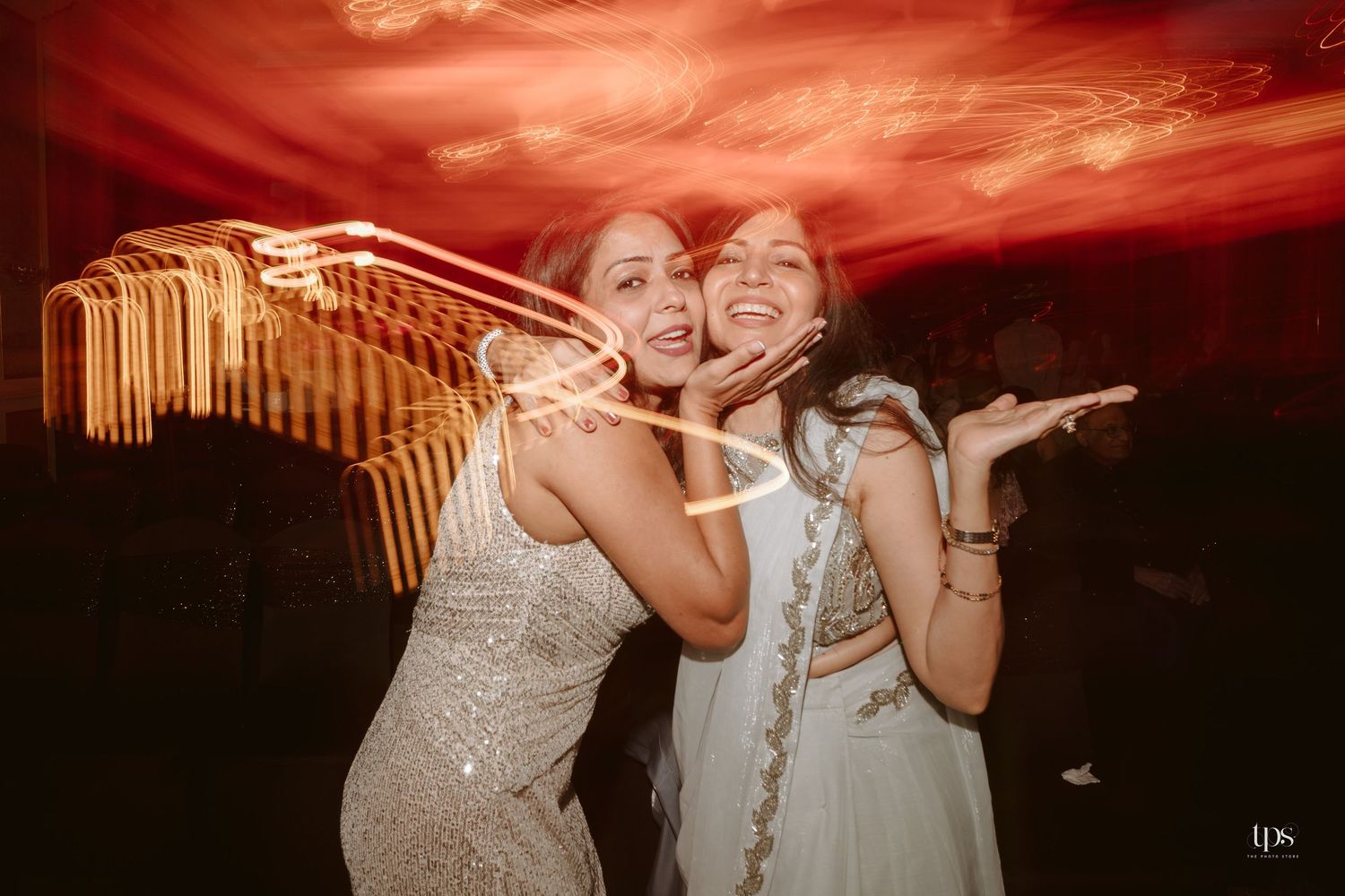 Two women in elegant attire smiling and posing at a festive party with colorful light trails in background.