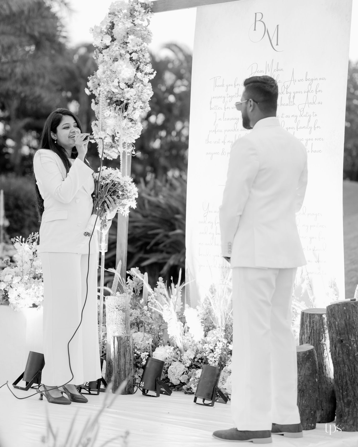 Black and white photo of a bride in a white suit speaking into a microphone beside a groom at an outdoor wedding ceremony with floral decor.