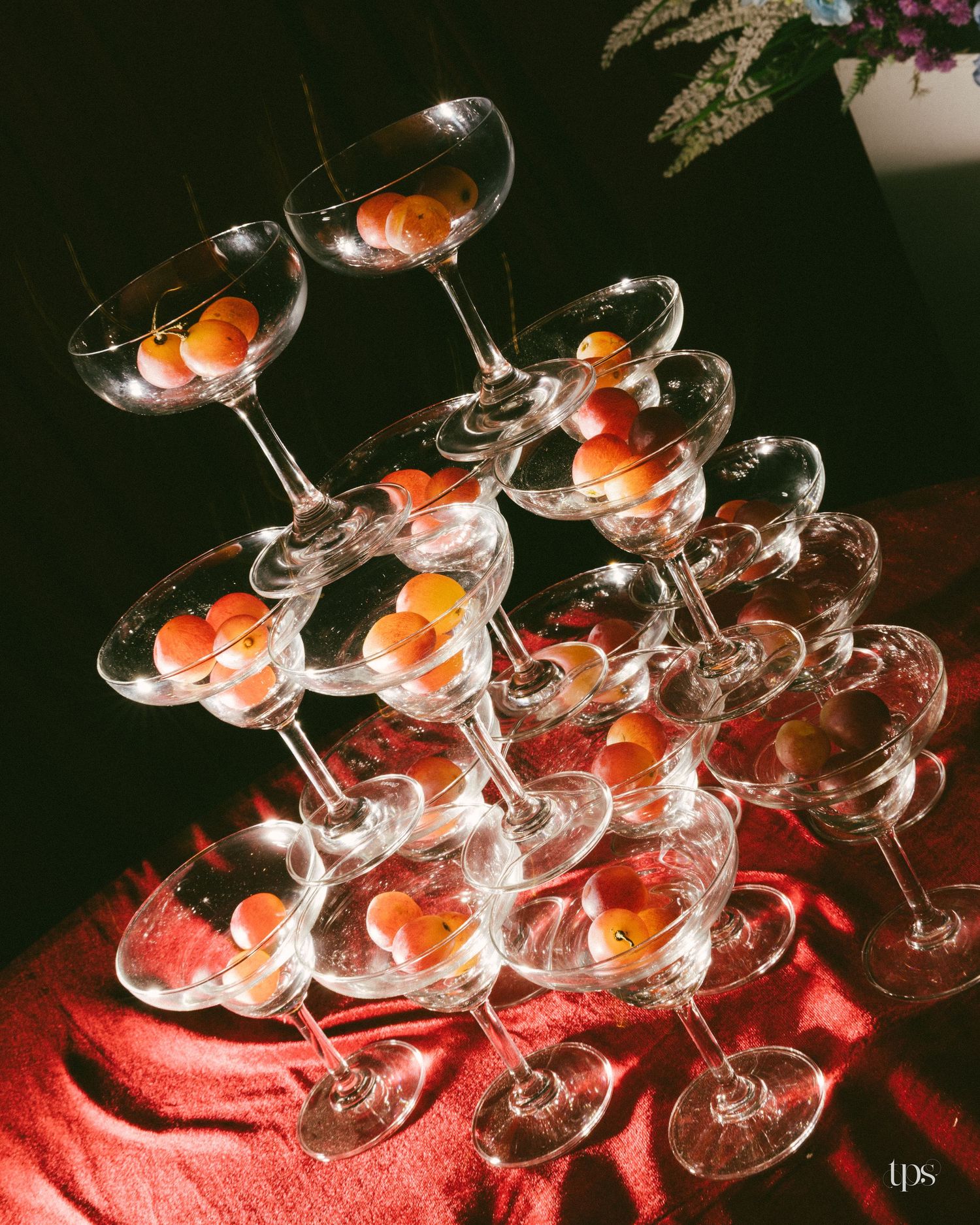 Stacked champagne coupe glasses forming a tower, each filled with small orange fruits, on a draped red cloth in dramatic sunlight.