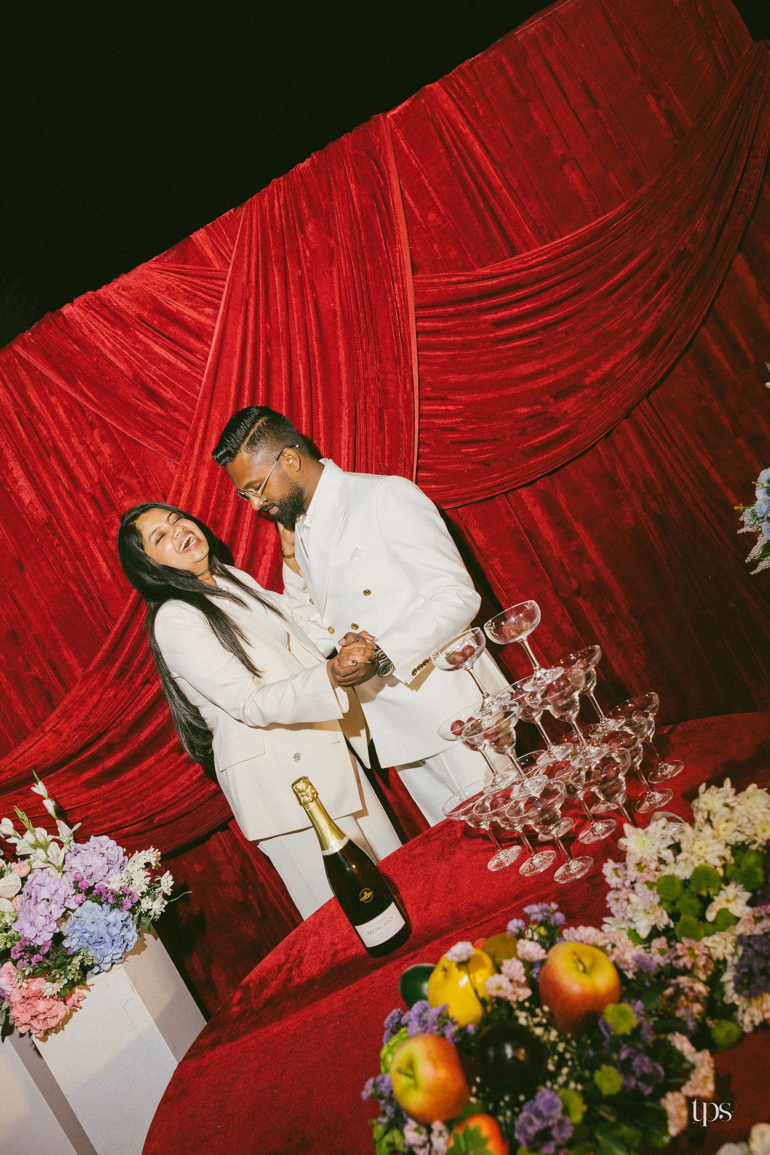 Couple in white outfits laughing and holding hands beside a champagne tower and bottle, with red velvet drapes and fresh flowers.