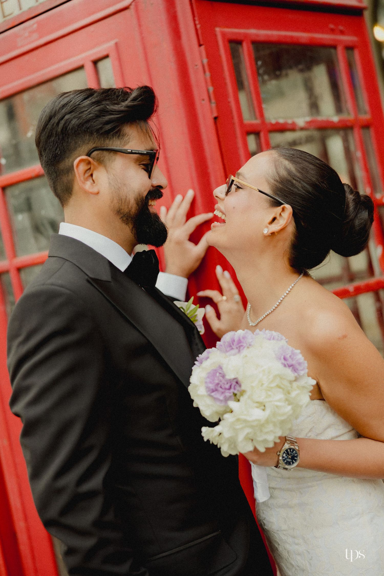 Bride and groom share a playful moment by a red phone booth, she holds white and purple flowers.