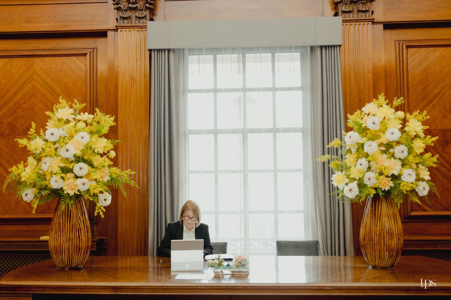 Woman seated at wooden desk flanked by large yellow and white floral arrangements in an elegant wood-paneled room.