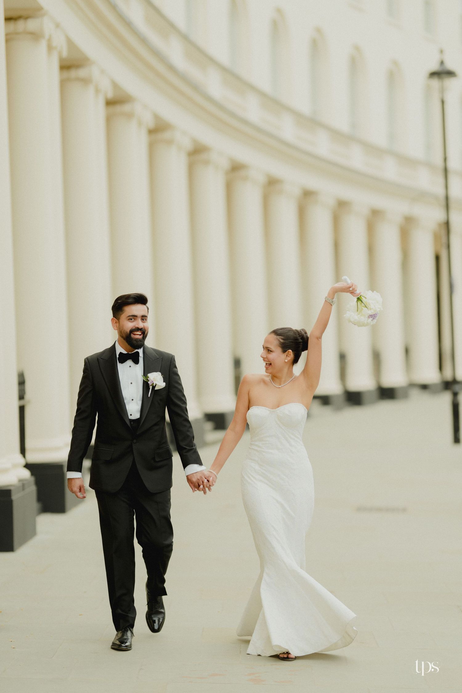 Bride in white gown raises bouquet while holding hands with groom in black tuxedo, walking past elegant white columns.
