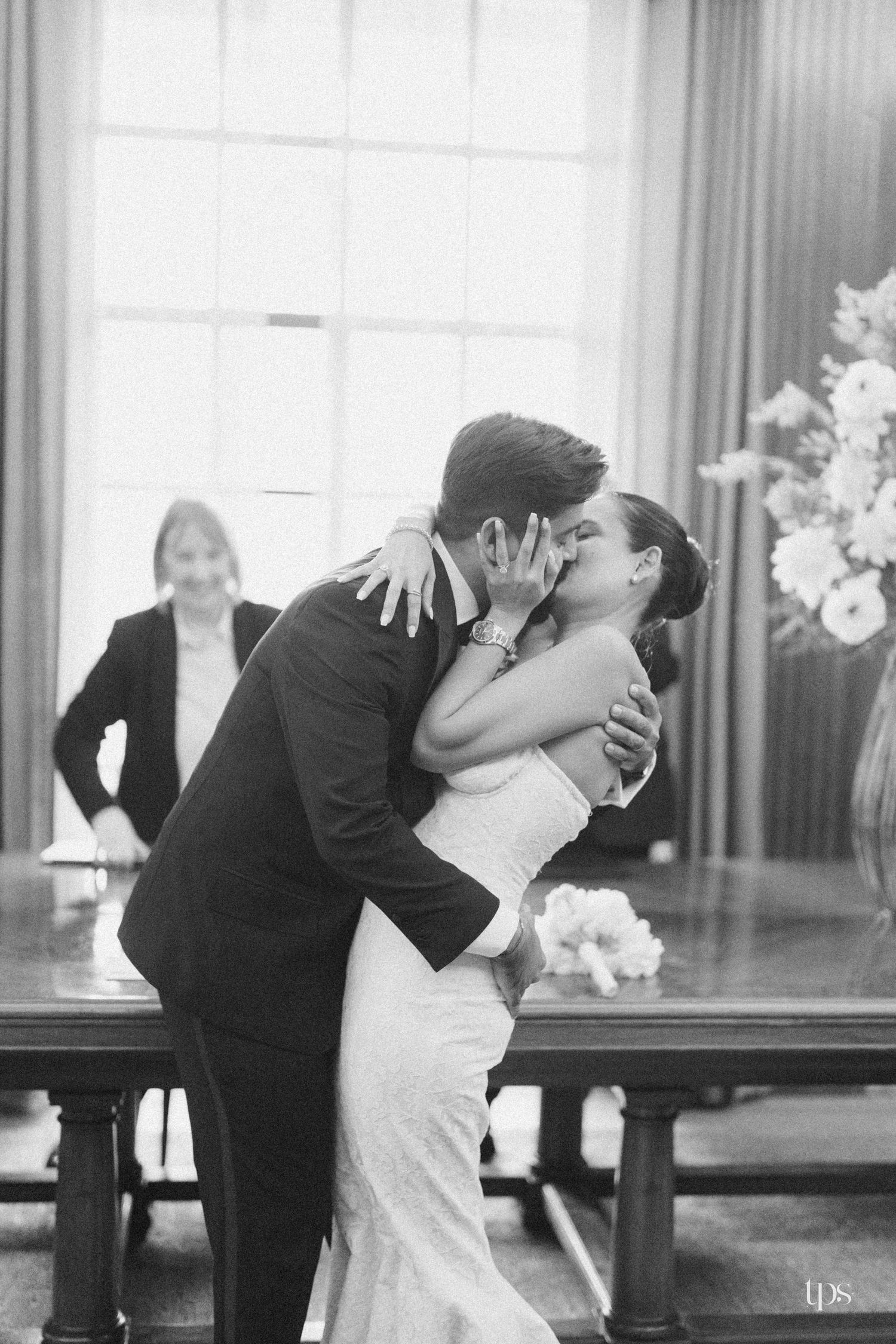 Black and white photo of a bride and groom sharing their first kiss at a wedding ceremony indoors.