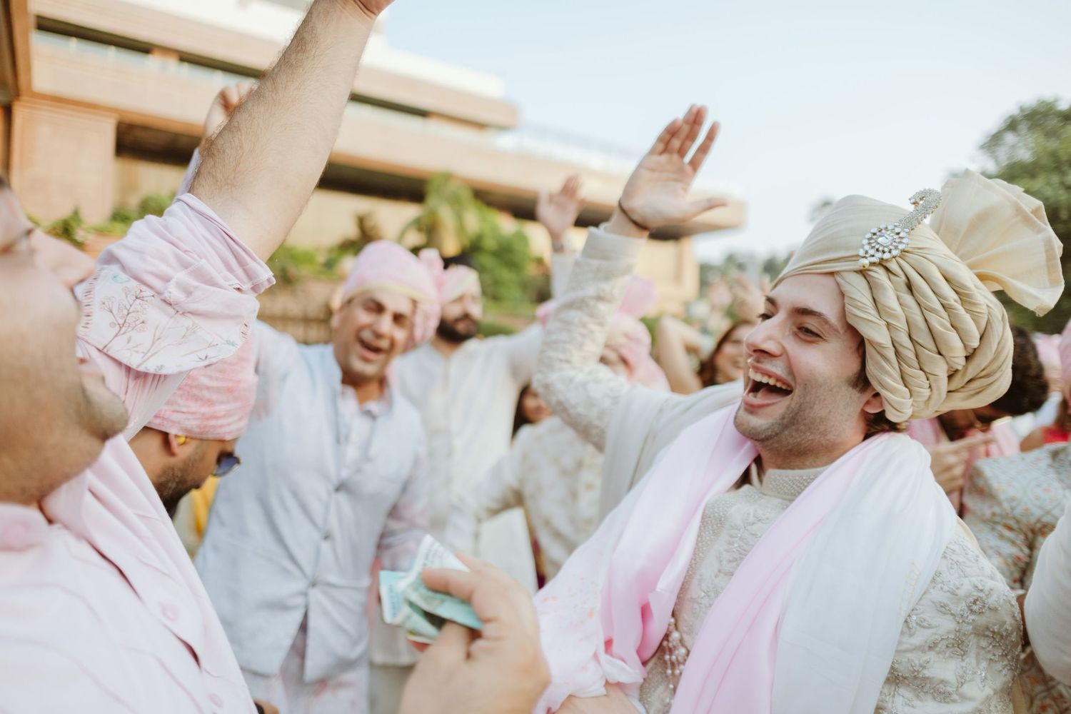 Joyful men in traditional Indian wedding attire, including turbans, celebrating outdoors with raised hands and laughter.