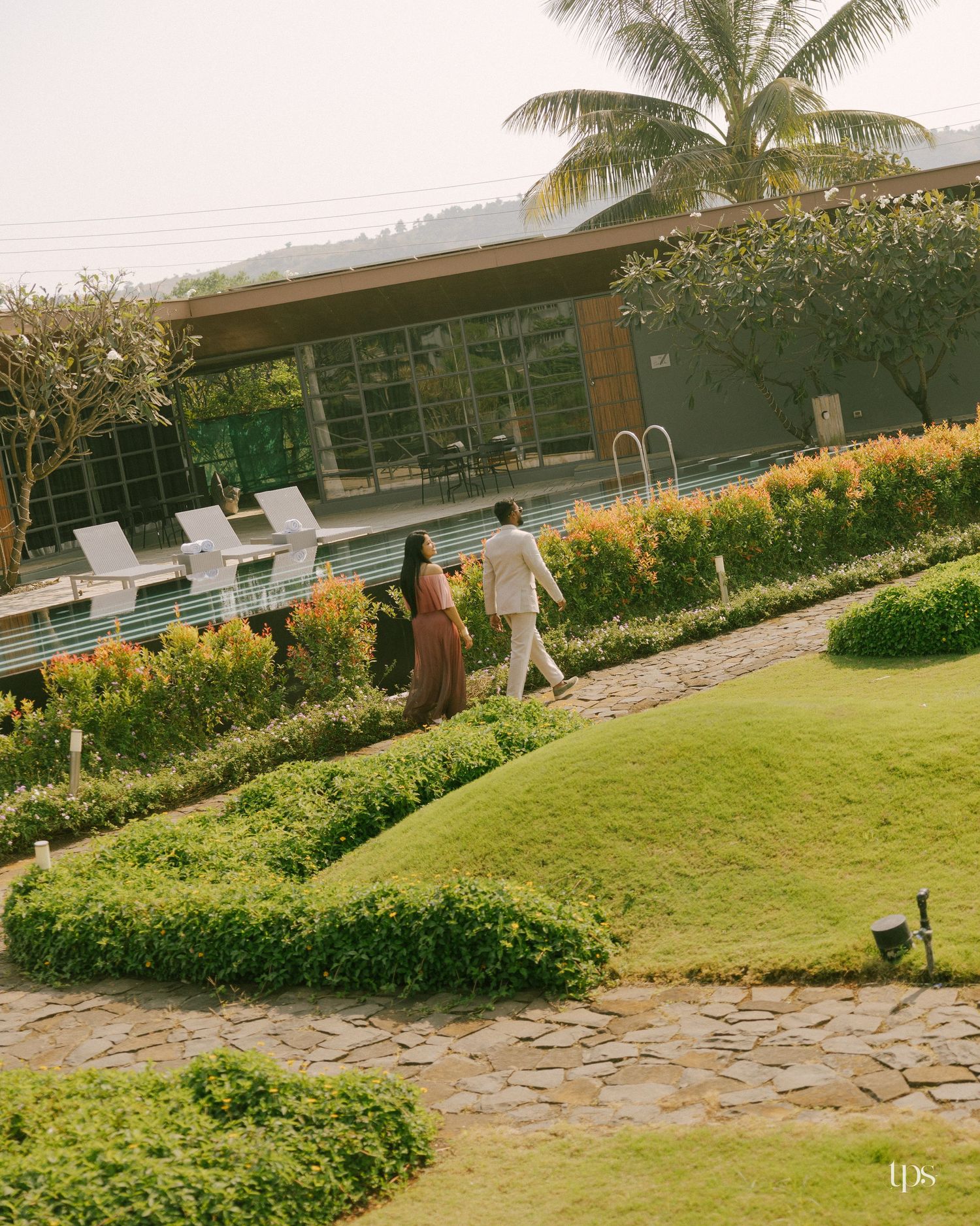 Couple walking along a stone path through a manicured garden with orange flowers, palm trees and a modern glass resort building.