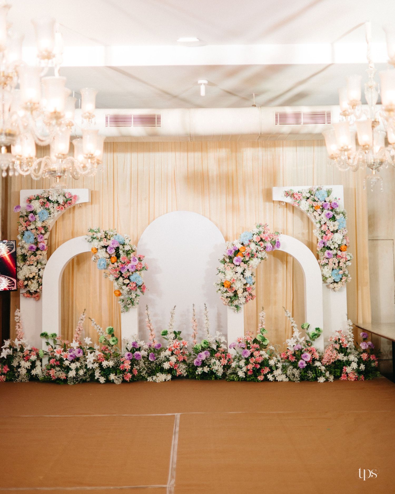 Elegant wedding stage with white arches, pastel floral arrangements, draped gold backdrop and crystal chandeliers.