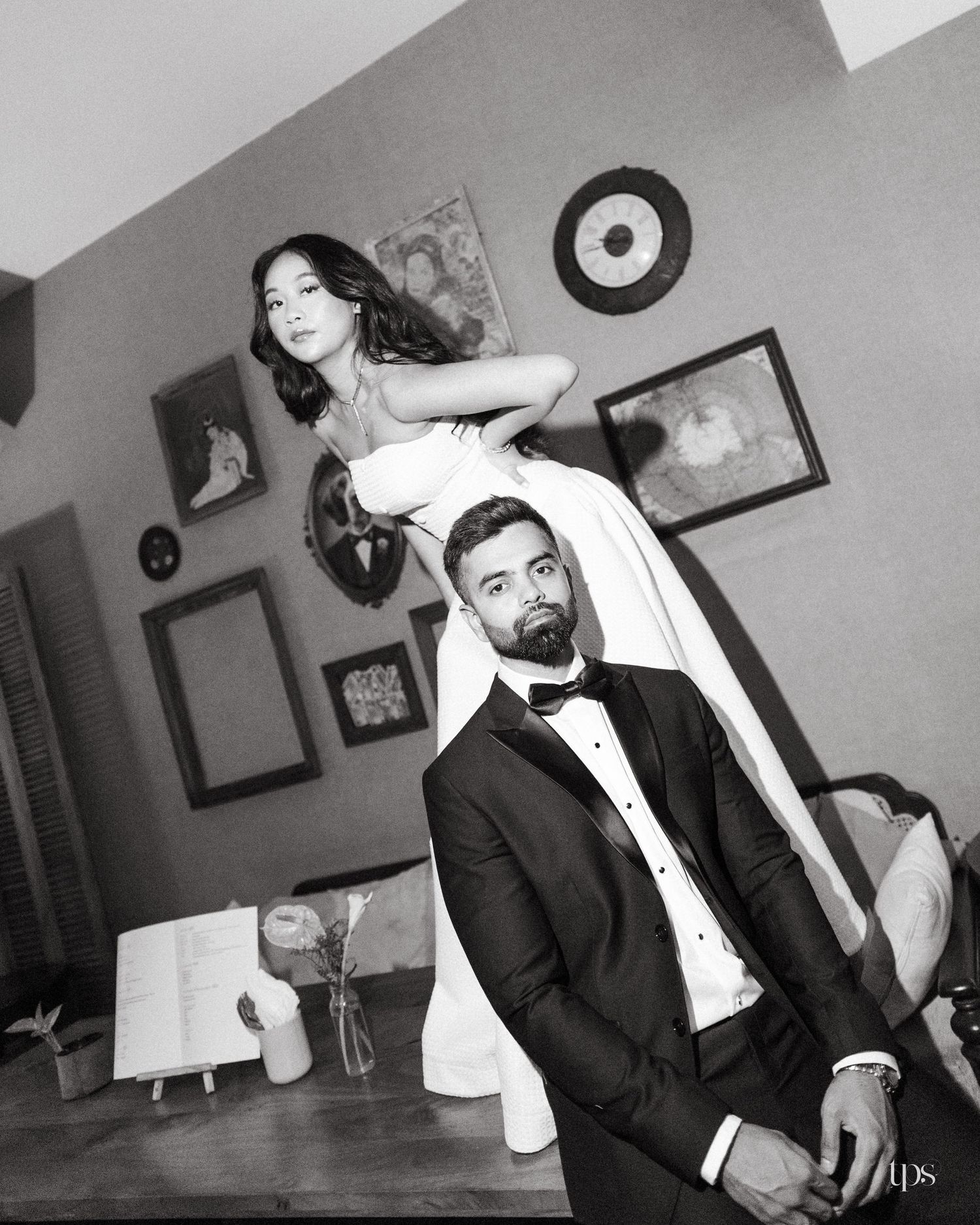 Black and white portrait of a bride in a white gown leaning over a seated groom in a tuxedo and bow tie in a vintage-style room.