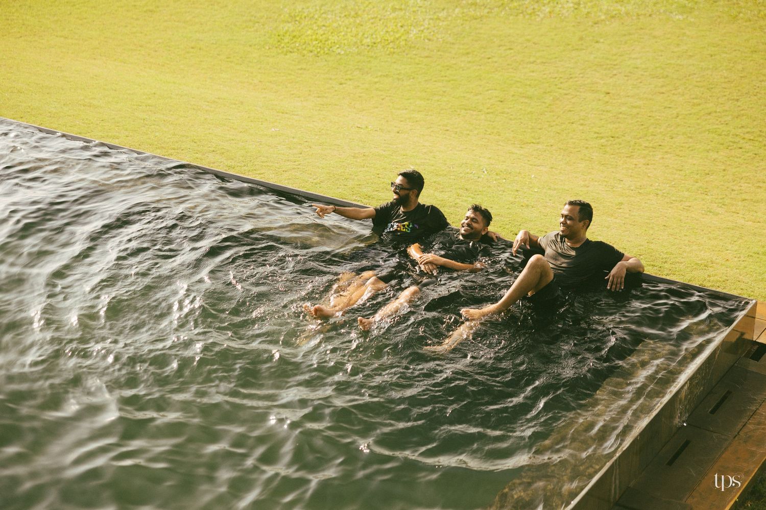 Three men in dark t-shirts relax in the water of an infinity pool at the edge of a lush green lawn.