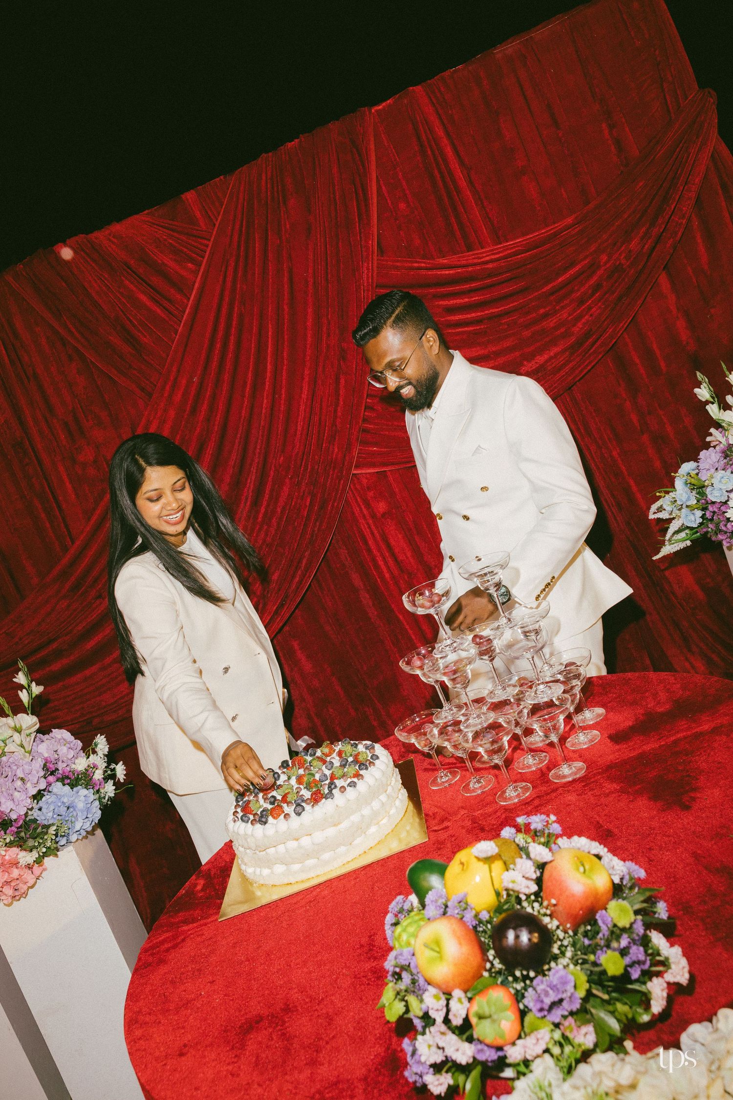 Couple in matching white outfits cutting a berry-topped cake beside a champagne tower on a red velvet table with fruit and flowers.