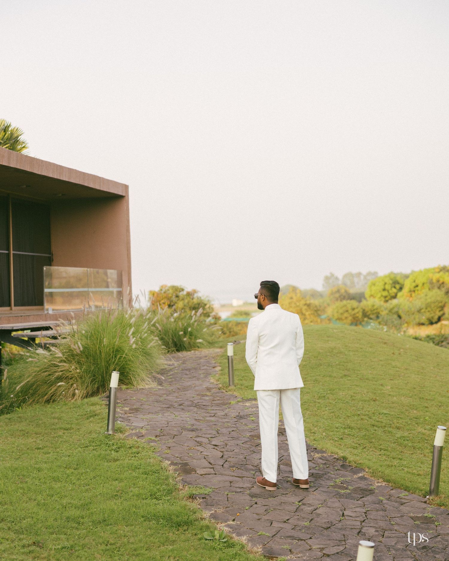 Man in a white suit stands on a stone pathway facing a modern building, surrounded by green lawns and shrubs under a hazy sky.