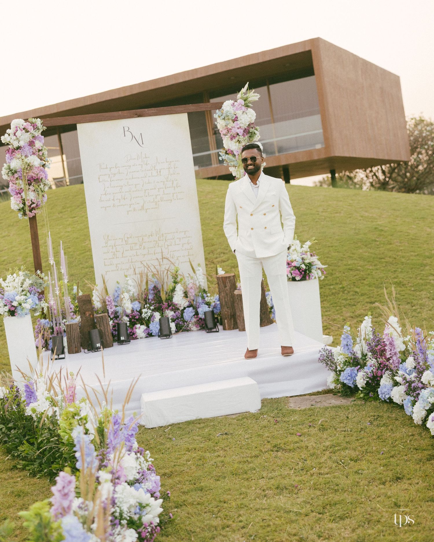 Man in a white suit stands on a white platform surrounded by purple and white flowers at an outdoor wedding ceremony setup on a green lawn.