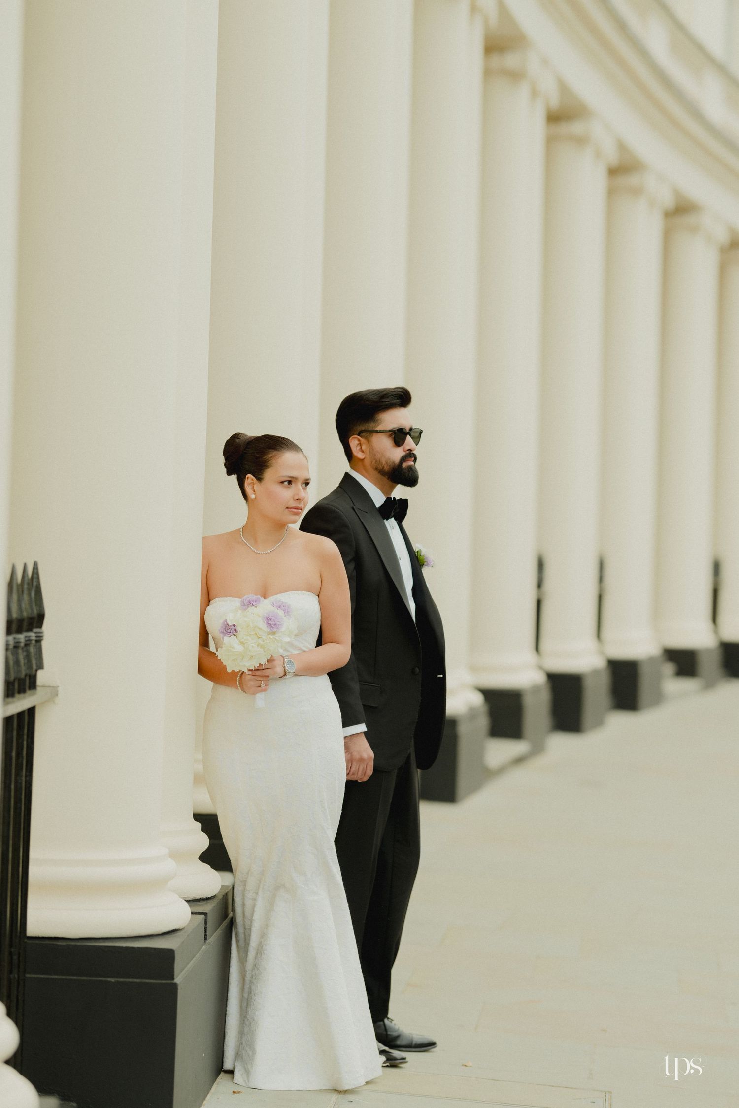 Bride in white strapless mermaid gown and groom in black tuxedo pose between grand white columns.