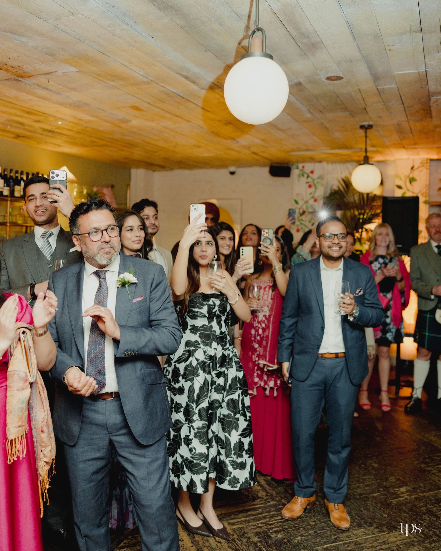 Wedding guests gather, smiling and recording on phones, dressed in formal attire in a warmly lit reception venue.