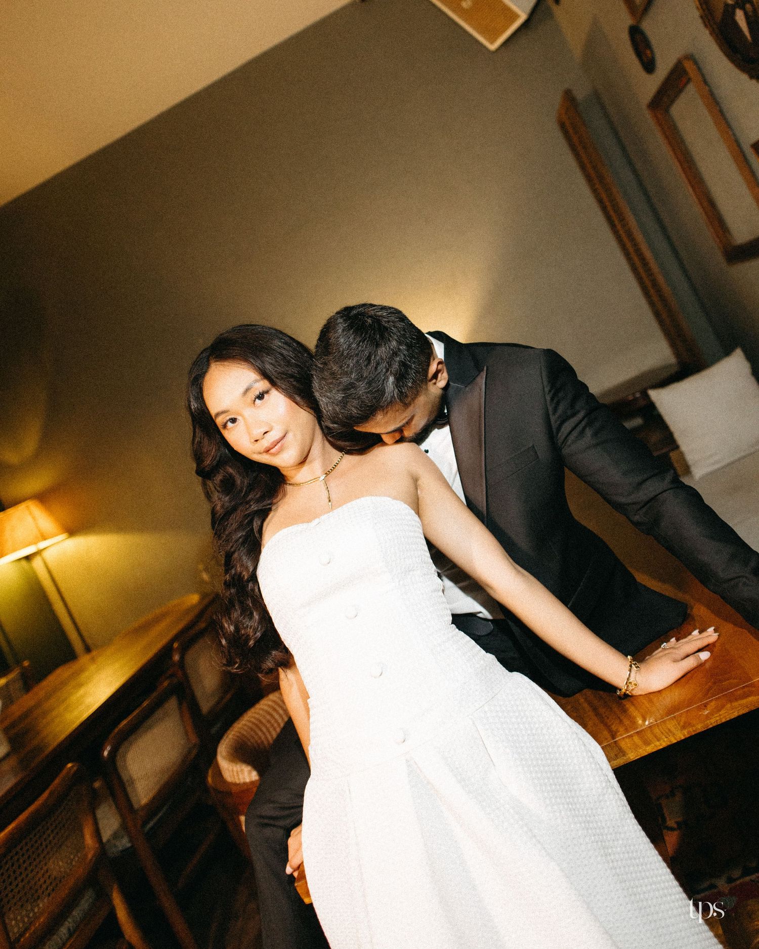A woman in a white strapless dress poses while a man in a black tuxedo kisses her cheek in a warmly lit, rustic room.