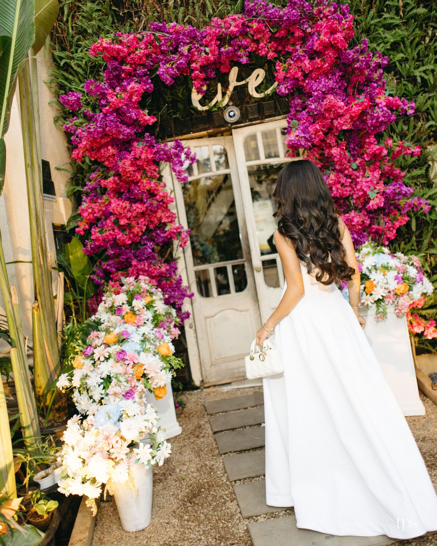 Bride in a white gown stands on a stone path facing a white door beneath an arch of pink and purple bougainvillea flowers.