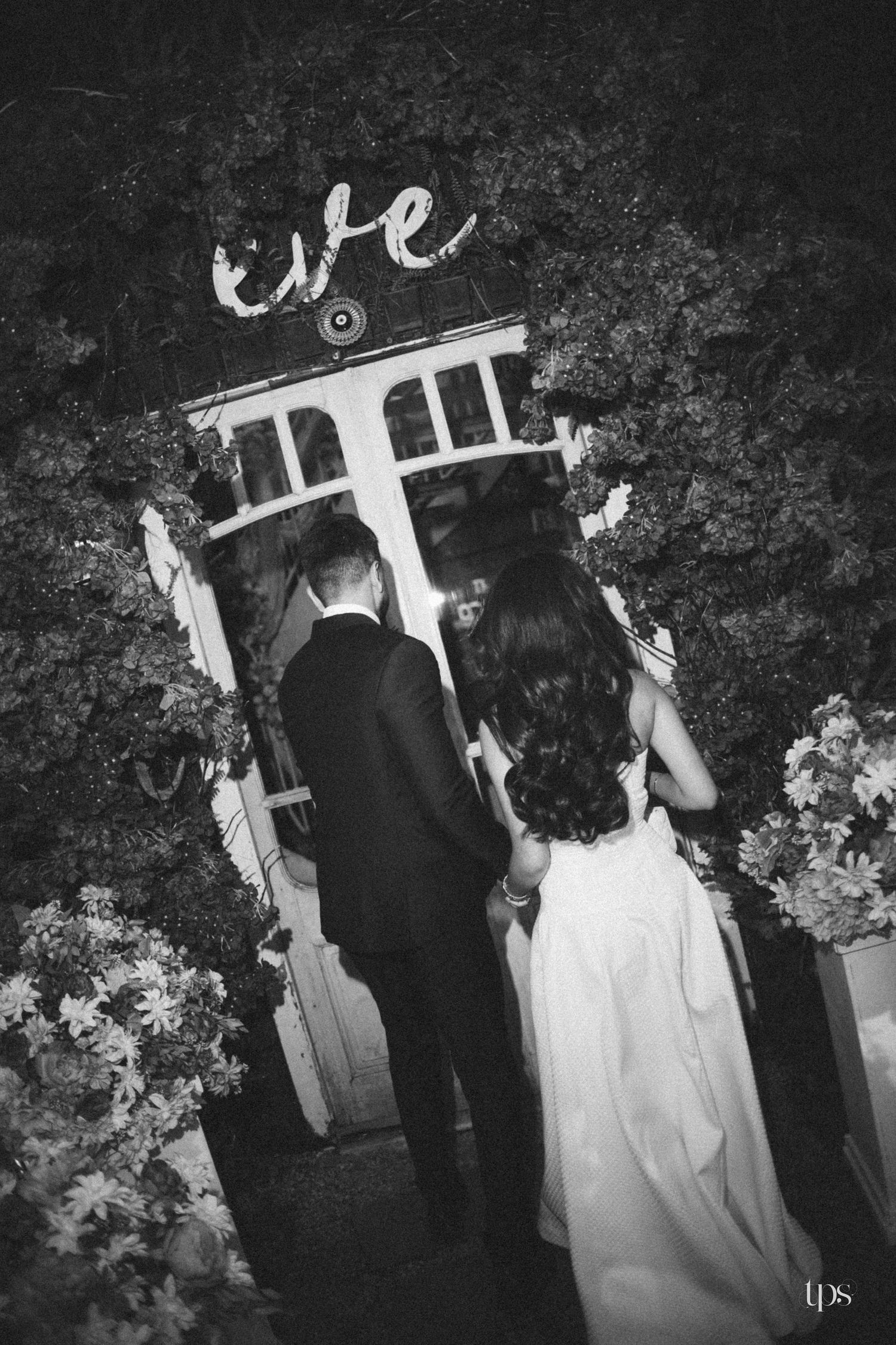 Black and white photo of a bride in a flowing gown and a groom in a suit standing at an ivy-covered doorway at night.