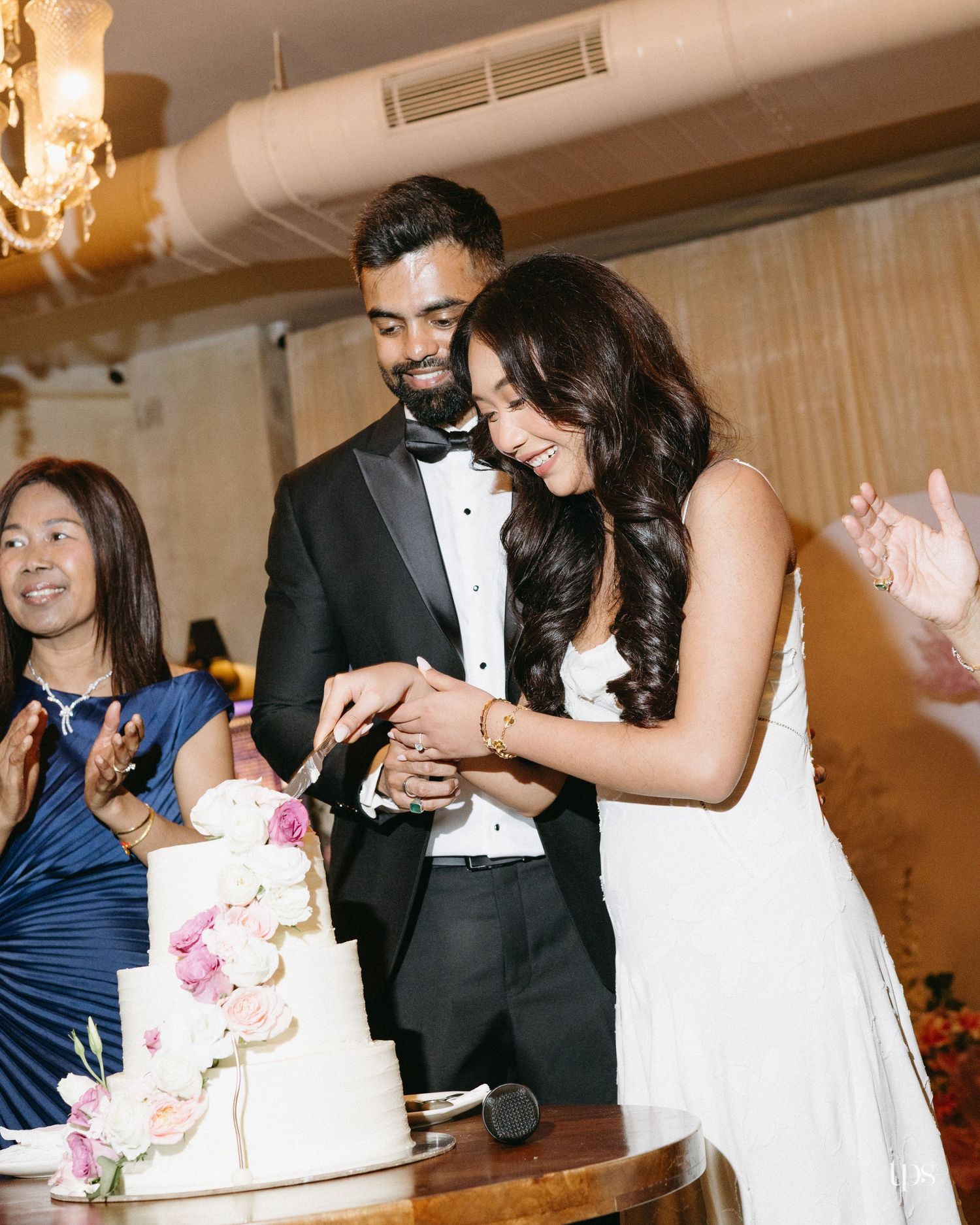 A bride in a white dress and a groom in a black tuxedo cut their floral-decorated wedding cake together while a guest smiles and claps.