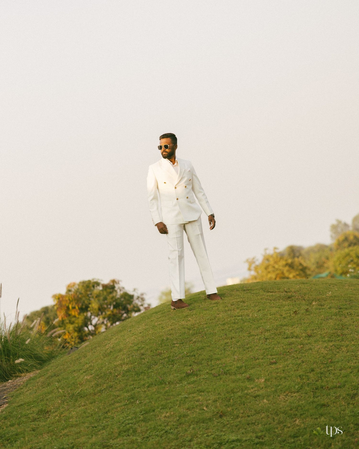 Man in sunglasses and a white double-breasted suit stands on a grassy hill with trees and a hazy sky behind him.