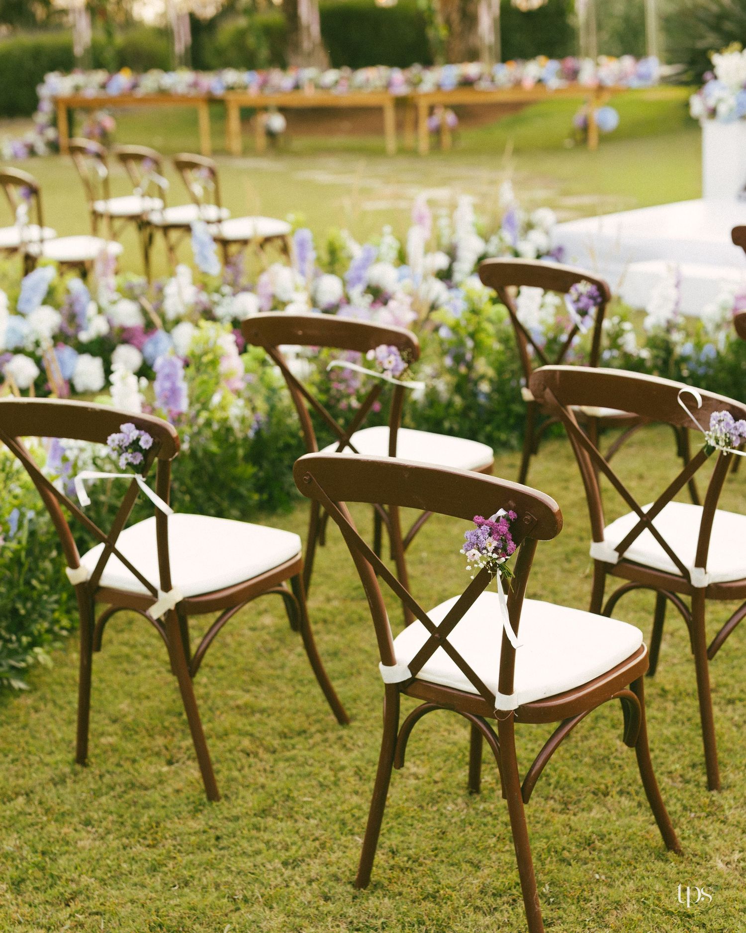 Wooden cross-back chairs with white cushions and floral sprigs line a grassy aisle at an outdoor garden wedding ceremony.