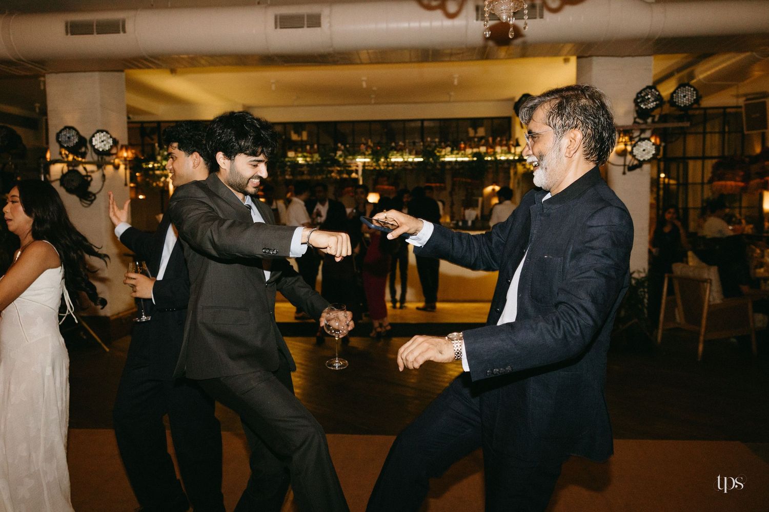Two men in dark suits dance energetically at a wedding reception while guests mingle in the warmly lit venue behind them.