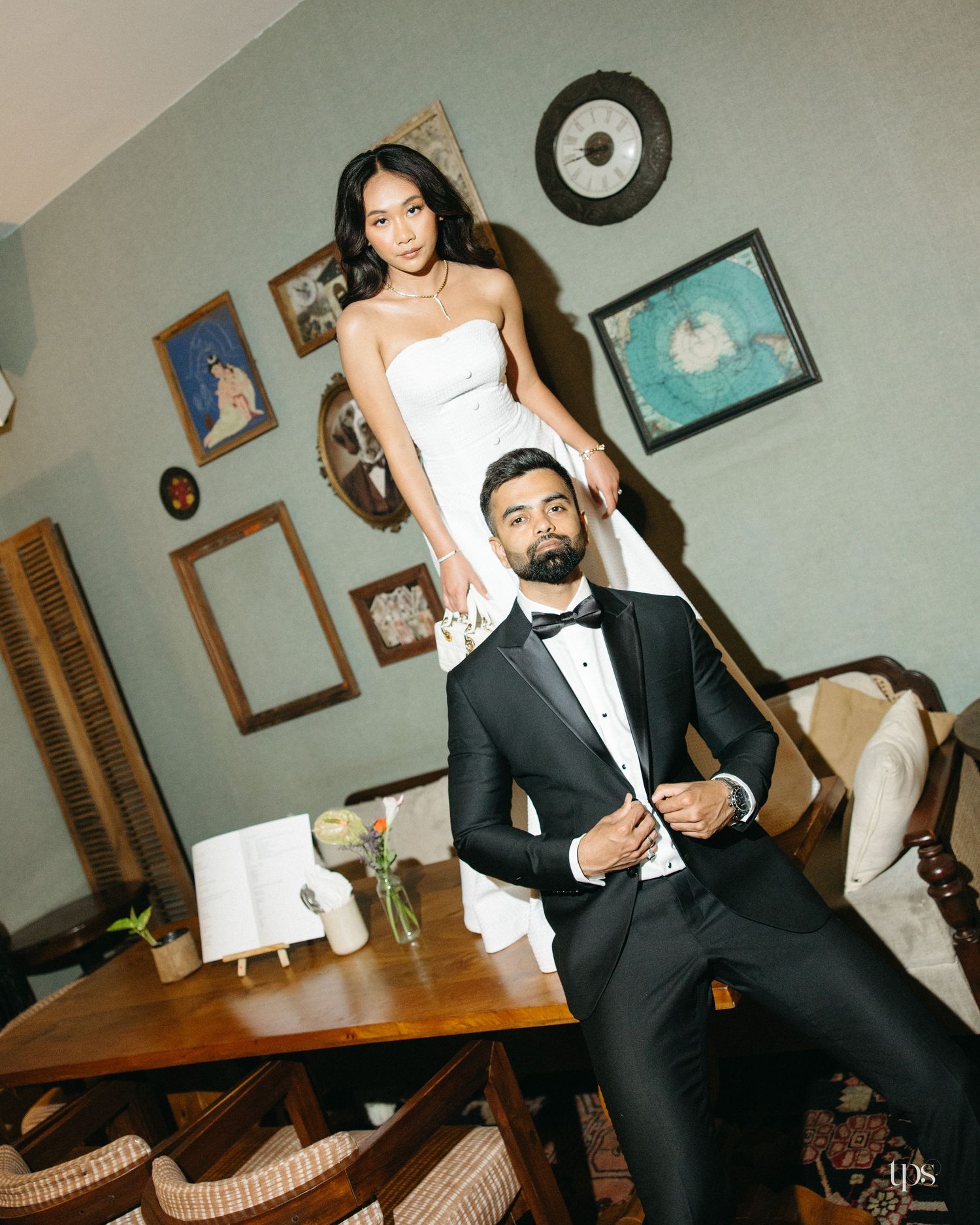 Bride in a white strapless gown stands above a groom in a black tuxedo and bow tie in a vintage room with framed art and a wooden table.