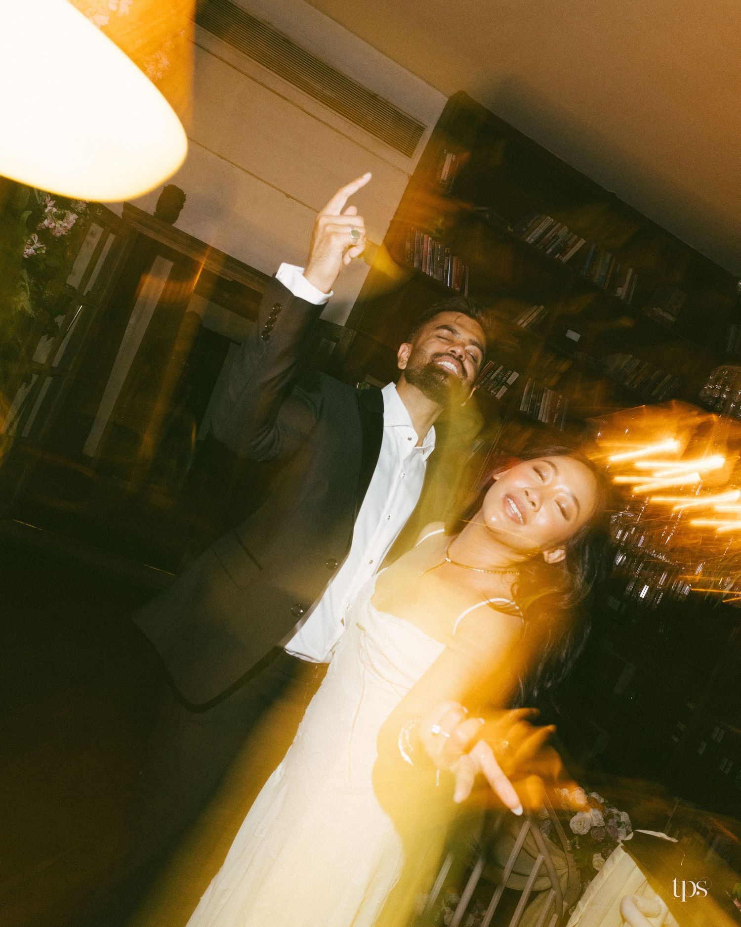 A smiling couple dances joyfully at an evening wedding reception, the man pointing upward as warm lights blur around them.