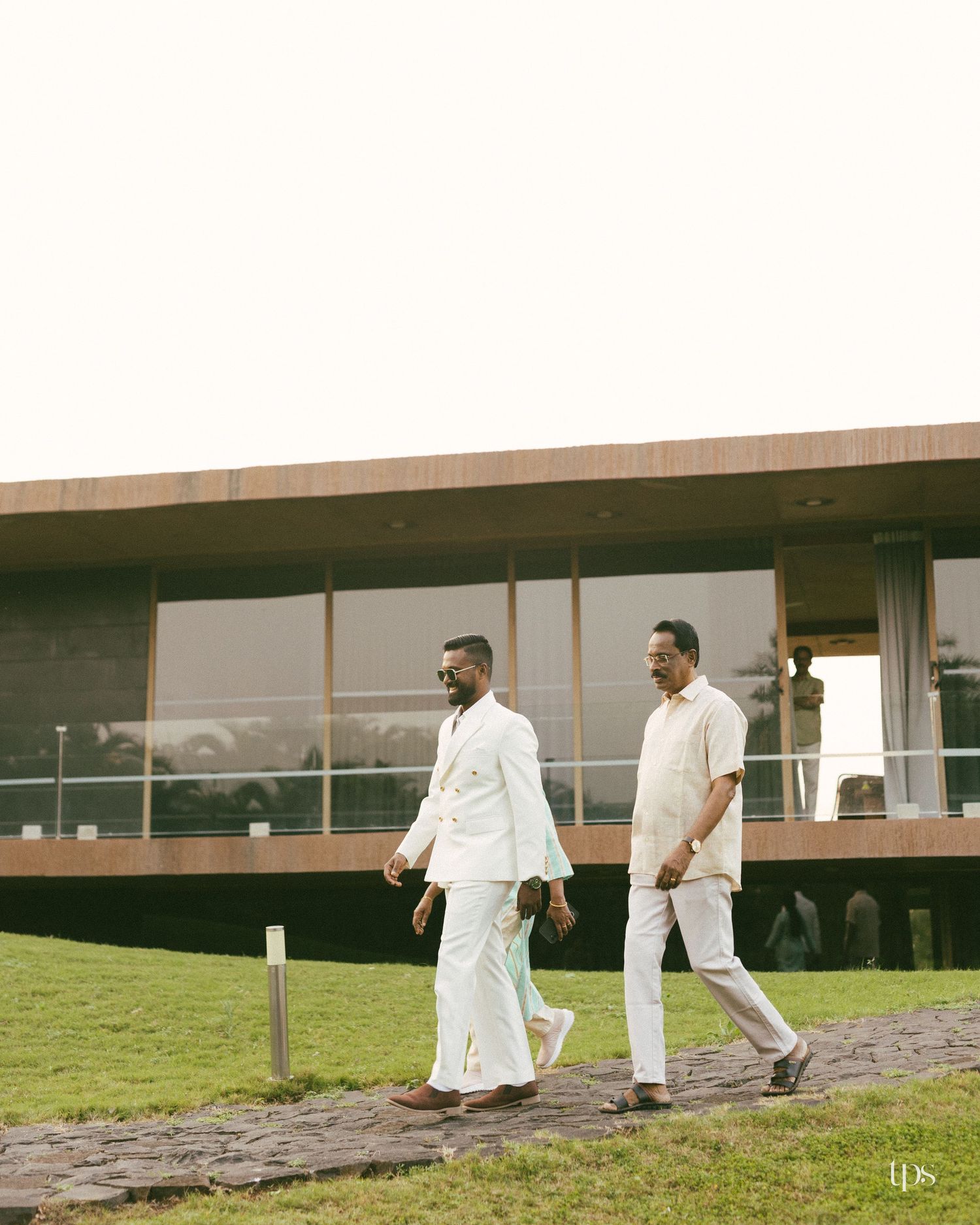 Two men in white linen outfits walk along a stone path on a grassy lawn toward a modern glass building at sunset.