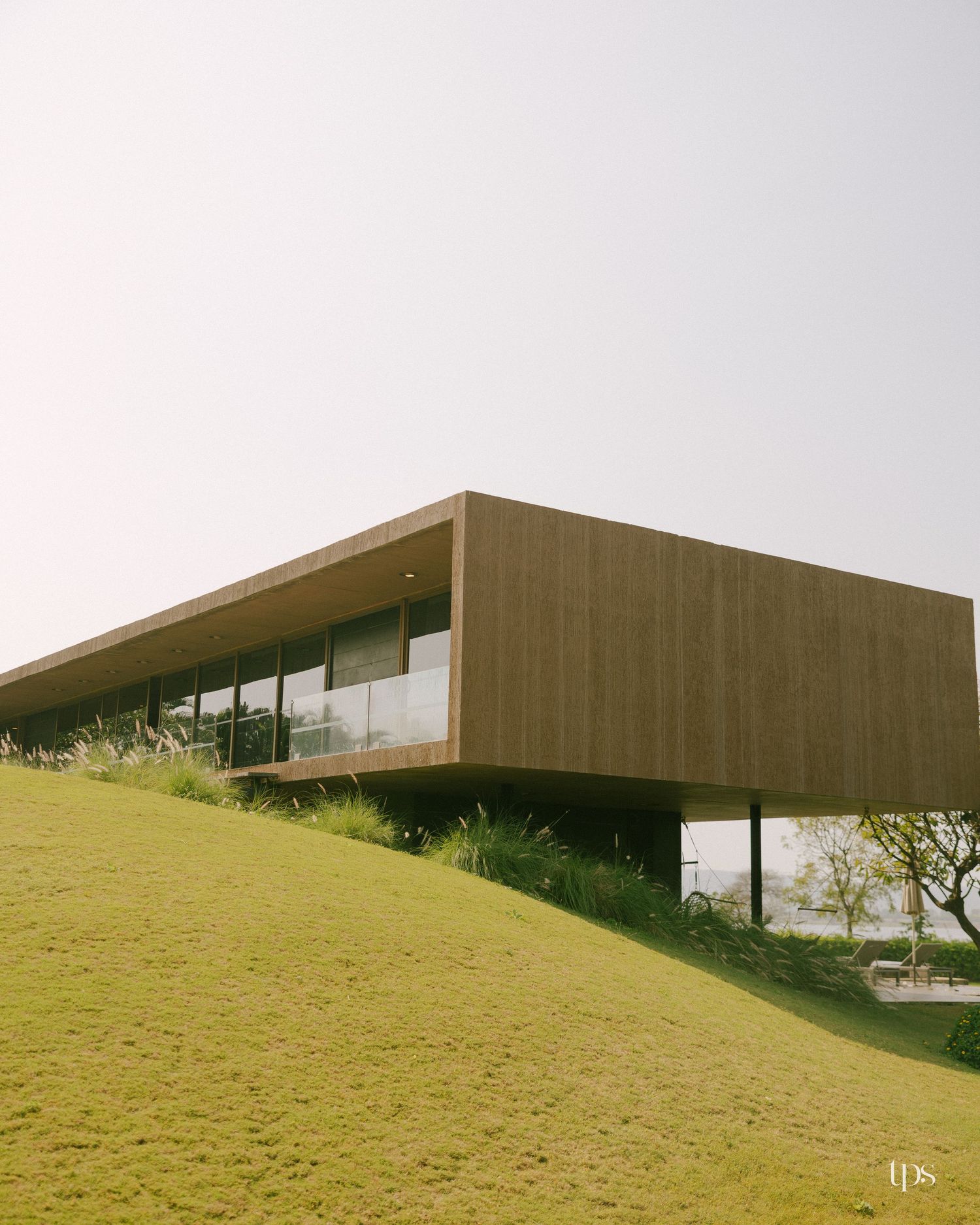 Modern wood-clad house with a cantilevered upper floor and glass walls rising above a sloping green grass lawn.