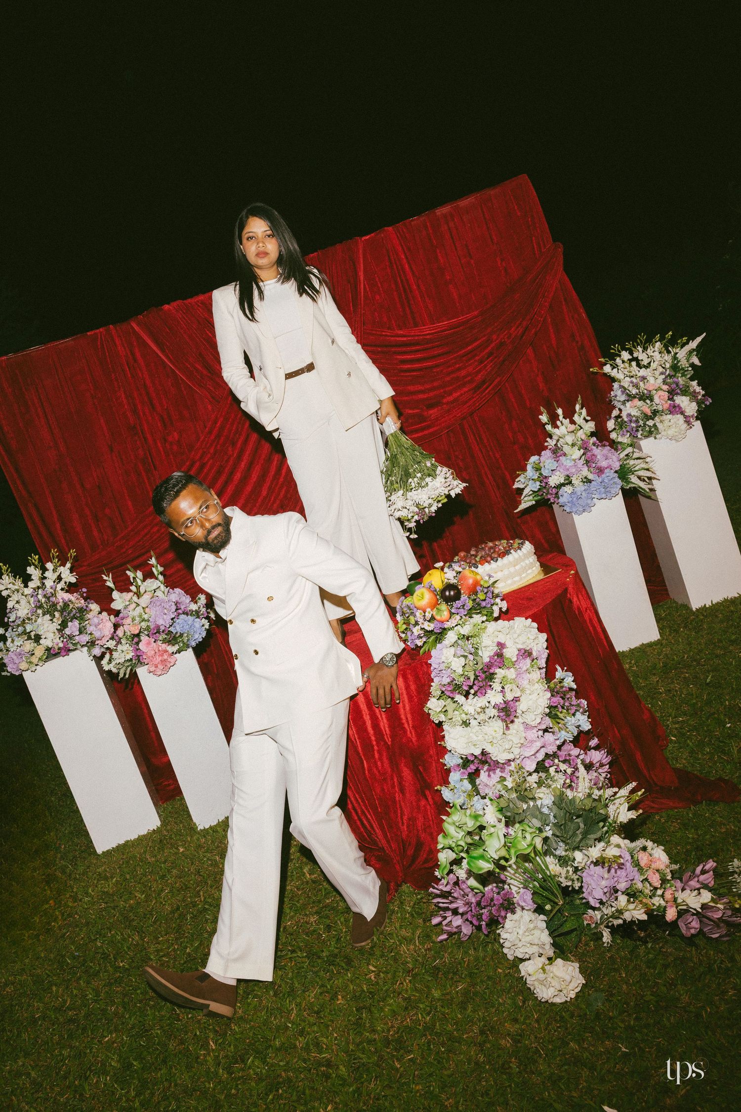 Couple in matching white suits pose outdoors at night with a red velvet drape backdrop, pedestals and lush pastel flower arrangements.