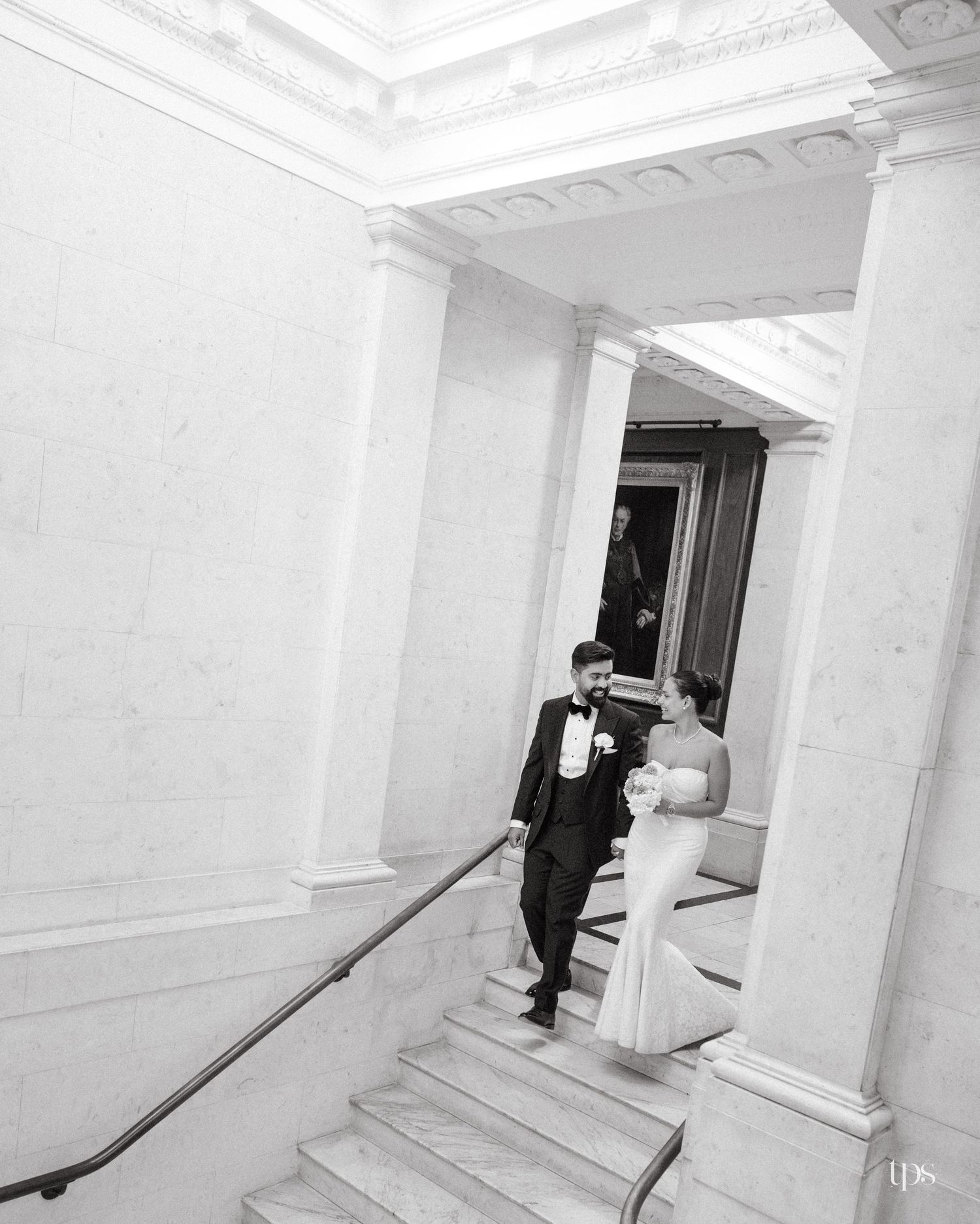 Black and white photo of a bride and groom descending elegant marble stairs with tall columns at a wedding venue.