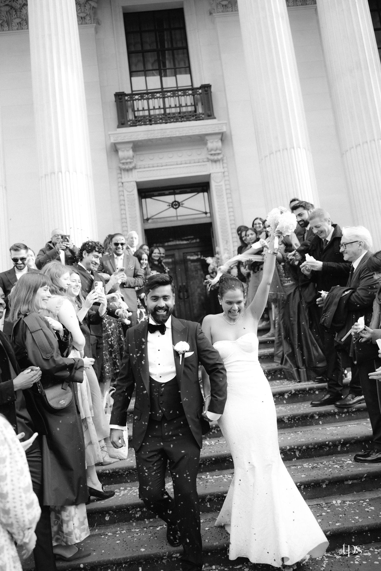 Black and white photo of a joyful newlywed couple exiting a grand church amid confetti, surrounded by cheering guests.