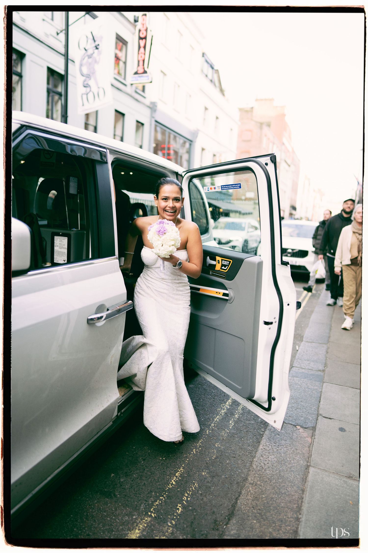 Smiling bride in white mermaid gown holding pink bouquet stepping out of a black cab on a city street.