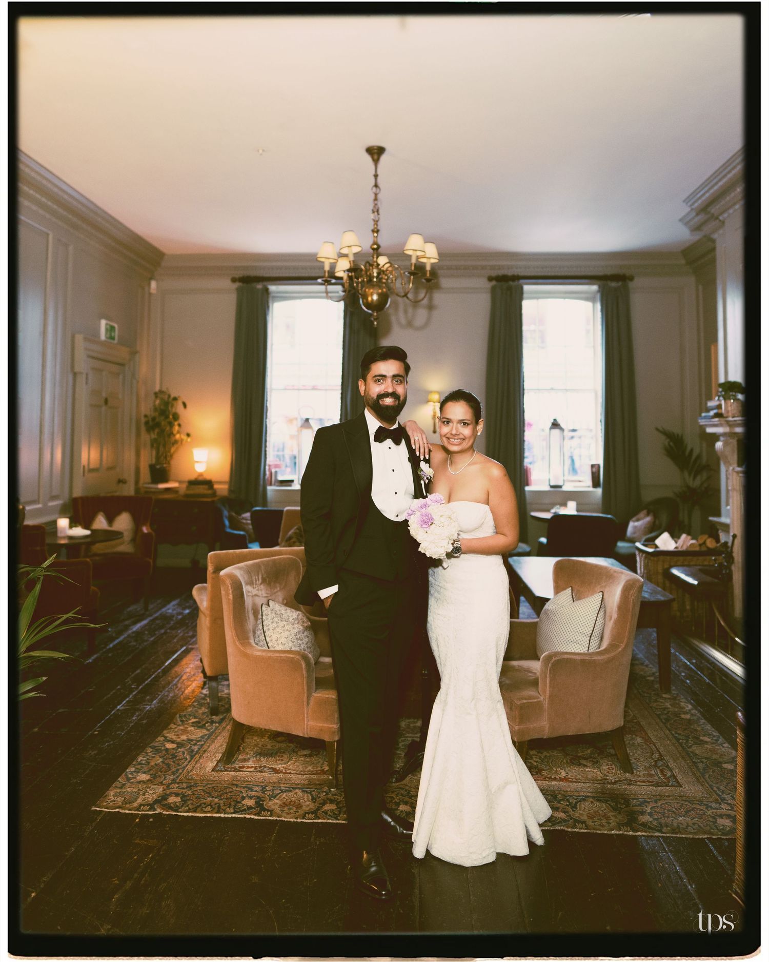 Bride in white mermaid gown and groom in black tuxedo pose together in an elegant chandelier-lit reception room.