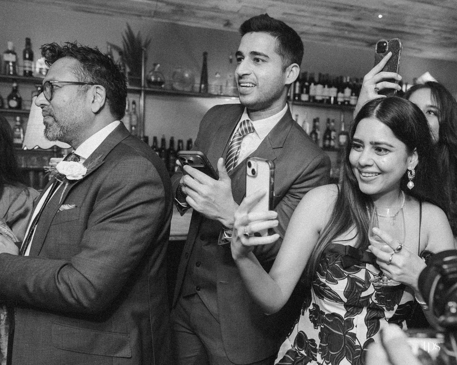 Black and white photo of elegantly dressed guests smiling and using phones at a bar during a wedding reception.