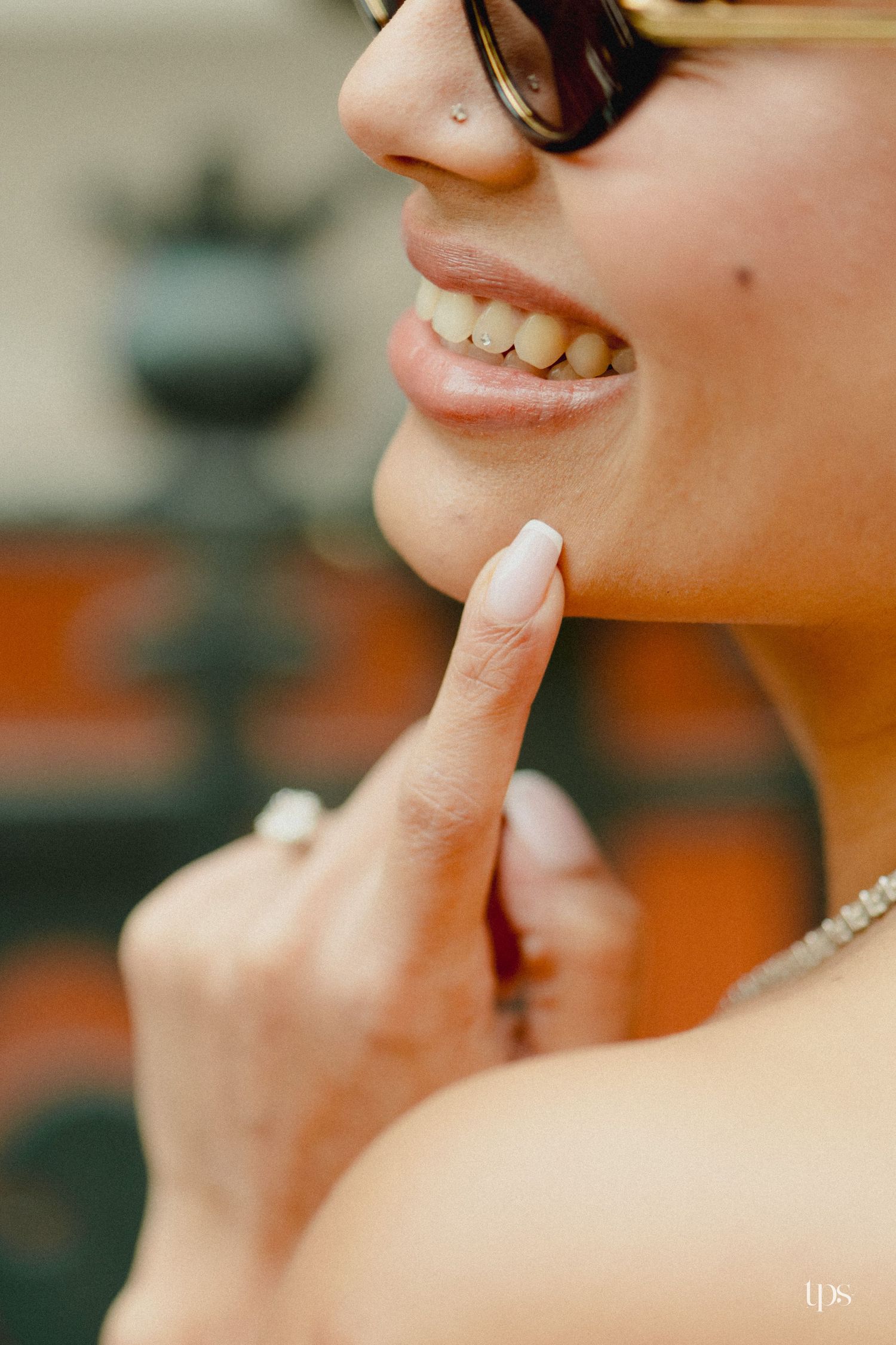 Close-up of a smiling woman with sunglasses, nose piercing, and manicured nails touching her chin.