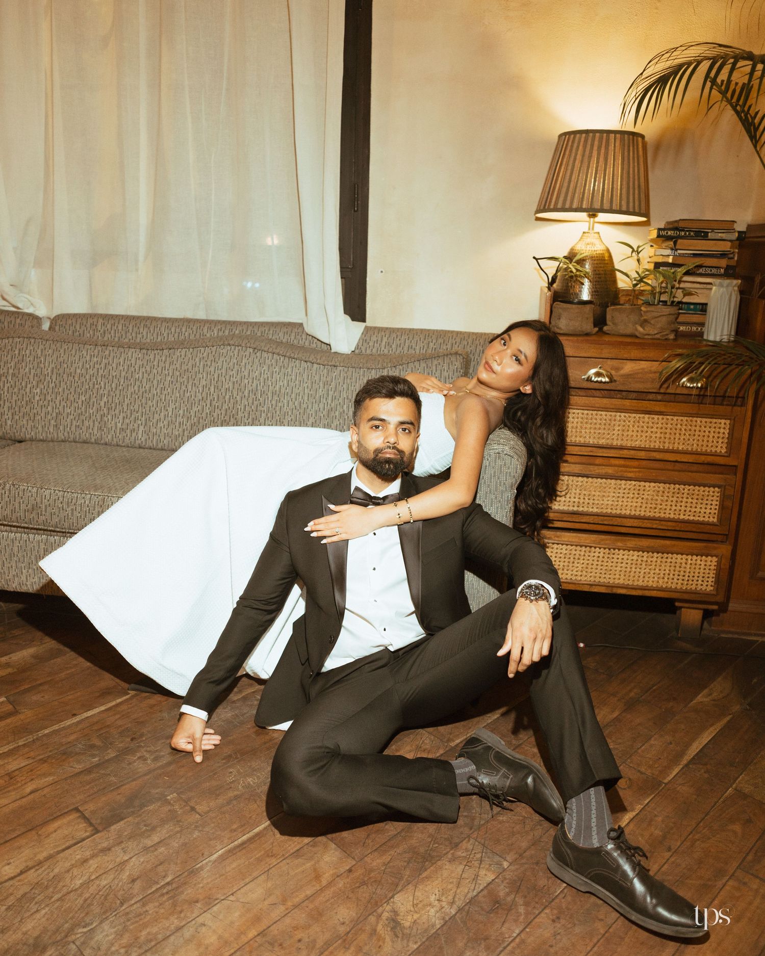 Couple in formal wear pose on a wooden floor, the man in a black tuxedo seated while the woman in a white gown leans on him.