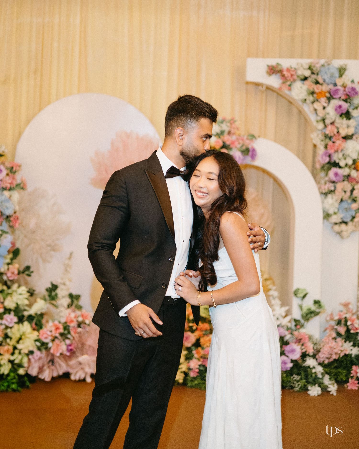 A man in a black tuxedo kisses a smiling woman in a white dress at a wedding with pastel floral arches and soft decor.