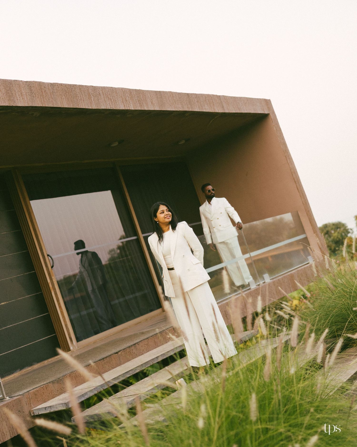 Couple in matching white suits pose on a modern rust-colored building with glass walls, surrounded by tall wild grasses.