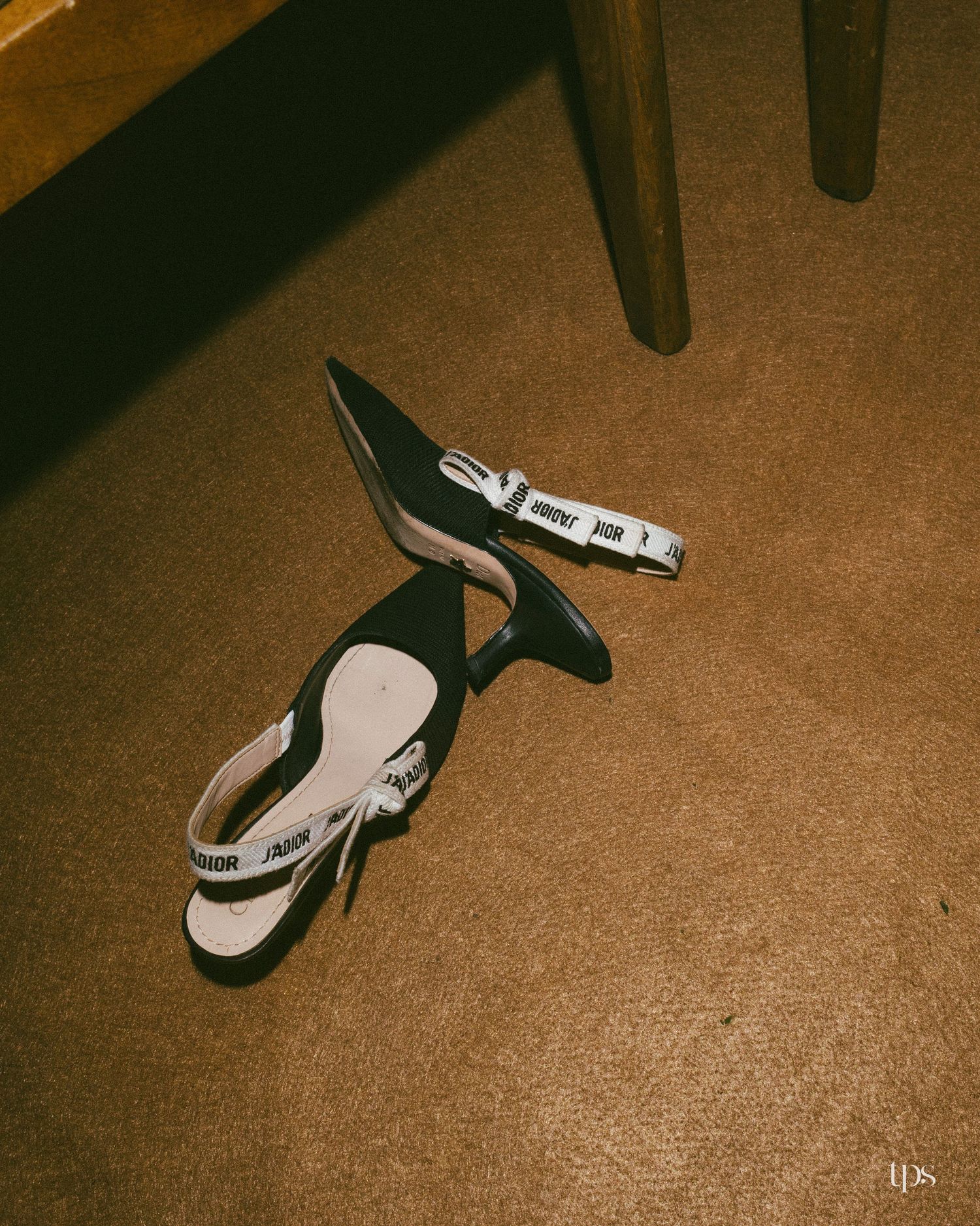 Pair of black pointed-toe slingback heels with white logo ribbon ankle straps lying on a brown carpet beside wooden chair legs.