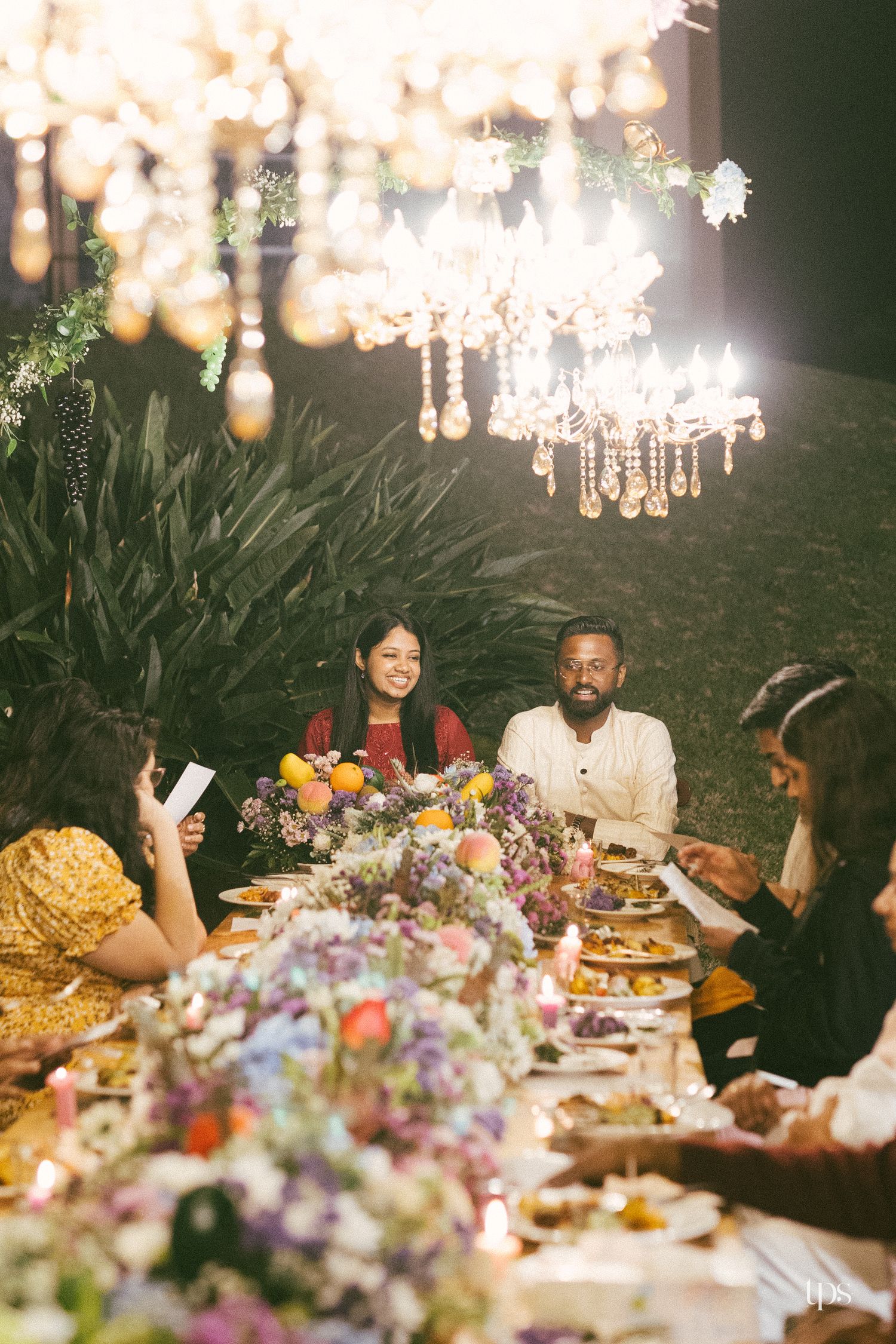 Guests enjoy an elegant outdoor dinner at a long table with floral centerpieces, glowing candles, and ornate crystal chandeliers overhead.